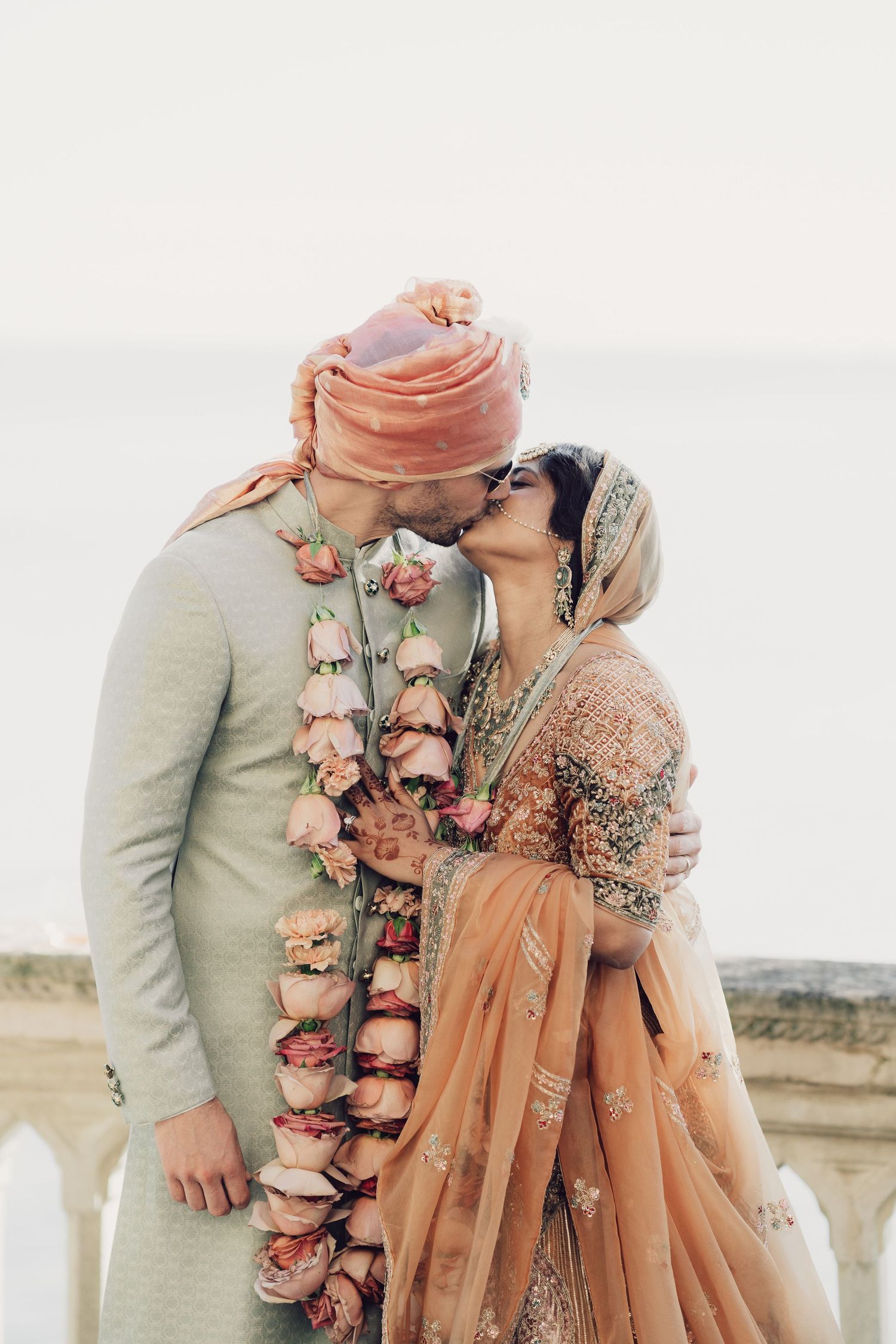 Indian bride and groom kissing wearing floral garlands and traditional wedding attire at outdoor ceremony.