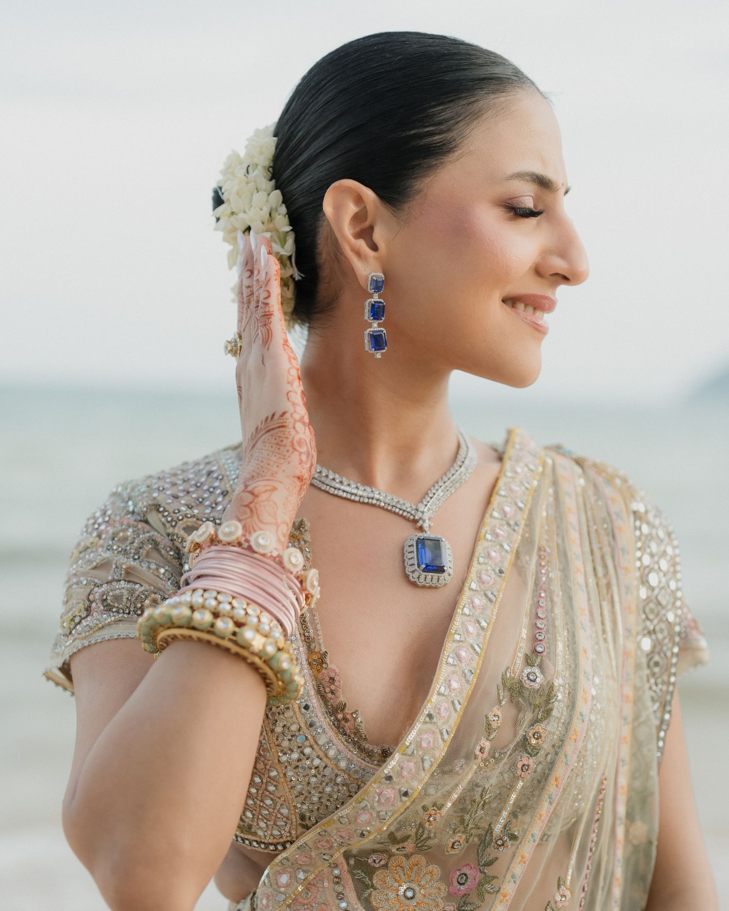 Indian bride in gold lehenga with sapphire necklace, mehndi hands, and floral hair accessory.