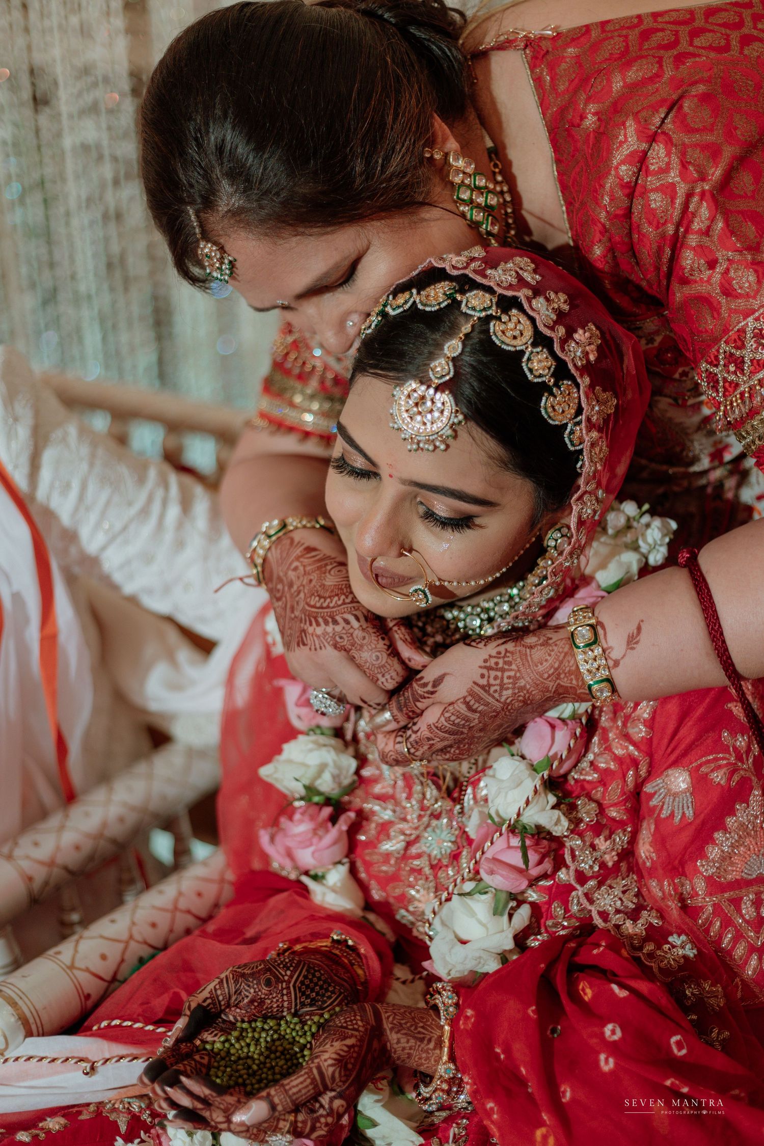 Bride in red bridal lehenga with mehndi hands holds white roses while being embraced during emotional wedding moment.