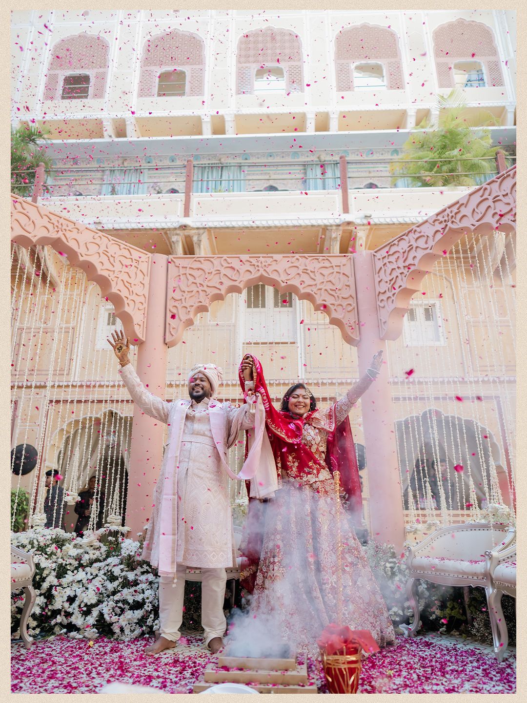 Indian bride and groom celebrate wedding ceremony under floral arch at ornate pink palace venue.