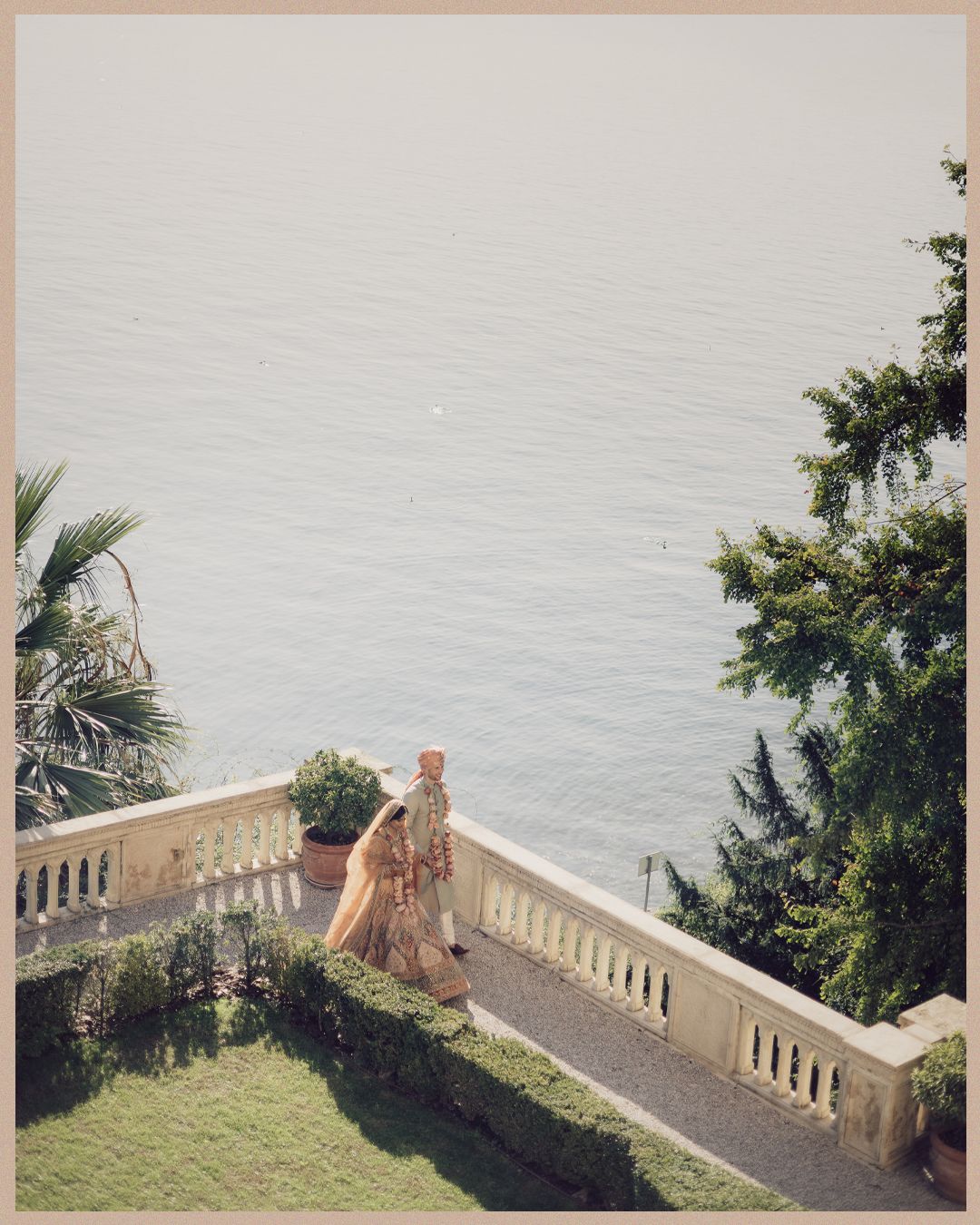 Indian wedding couple in traditional attire walk along elegant balcony terrace overlooking serene lake.