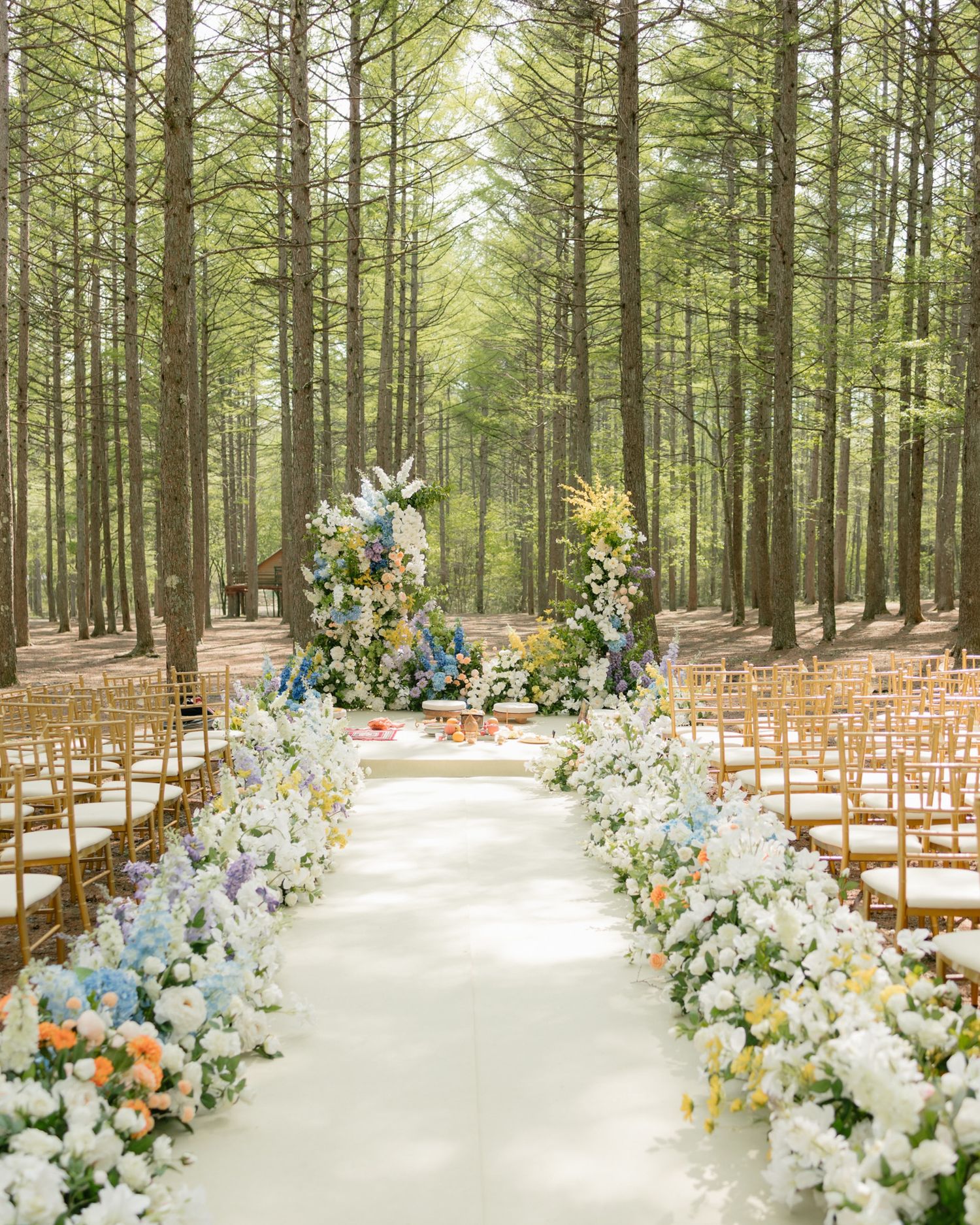 Enchanting outdoor forest wedding aisle lined with white and colorful flowers leading to a floral arch ceremony backdrop.