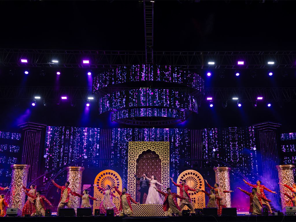 Elaborate wedding stage decorated with purple lights, floral curtains, and traditional dancers surrounding the couple.