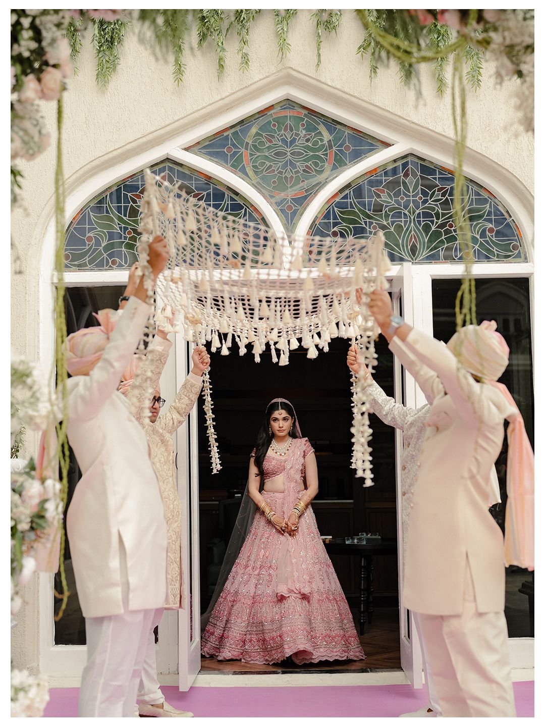 Indian bride in pink lehenga making a grand entrance under a floral canopy held by groomsmen.