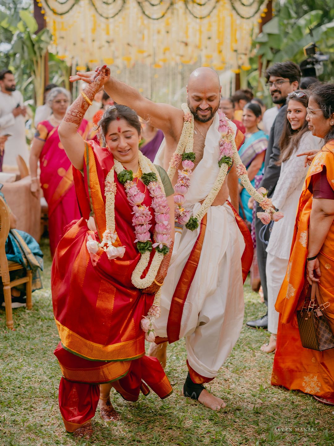 Bride in red saree and groom in white dhoti dance joyfully at outdoor South Indian wedding celebration.
