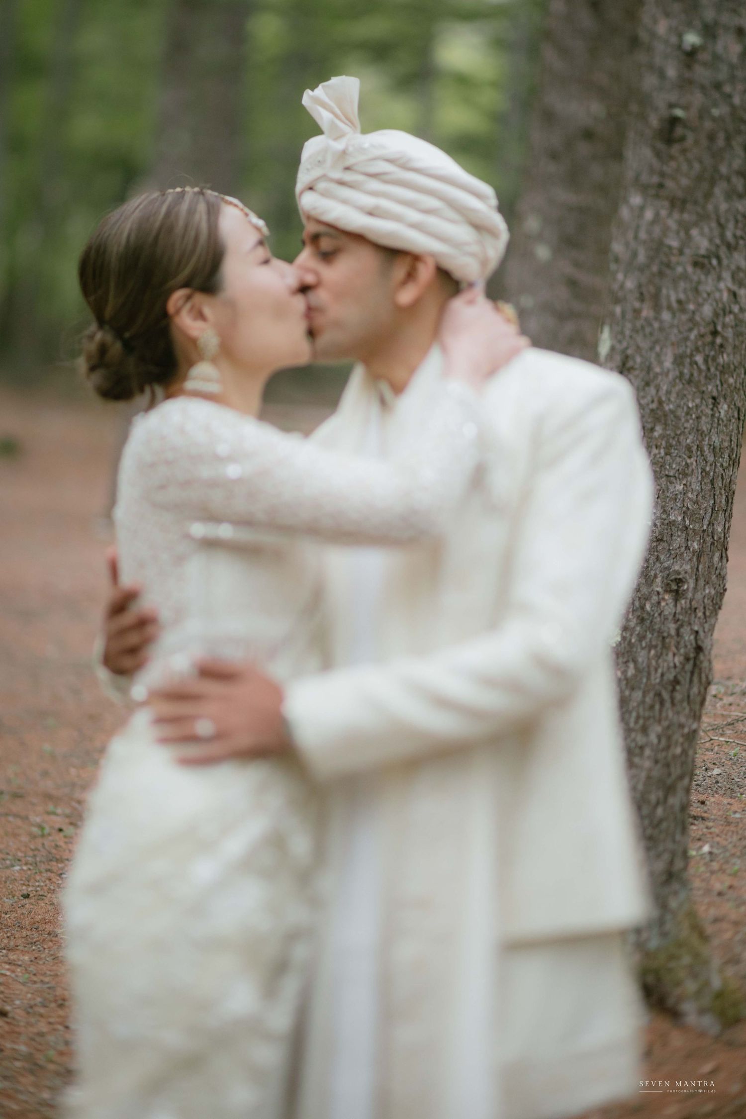 Newlyweds sharing first kiss in pine forest, groom wearing white sherwani and turban.
