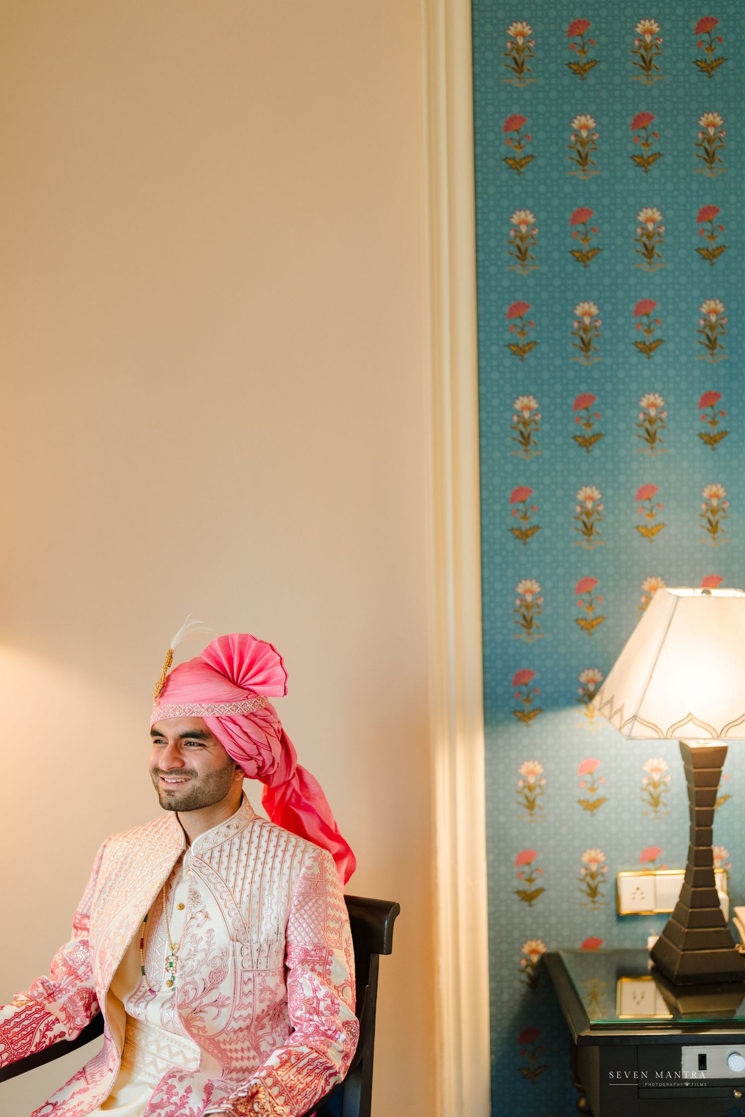 Handsome Indian groom in pink embroidered sherwani and turban poses in sunlit room with decorative wallpaper.
