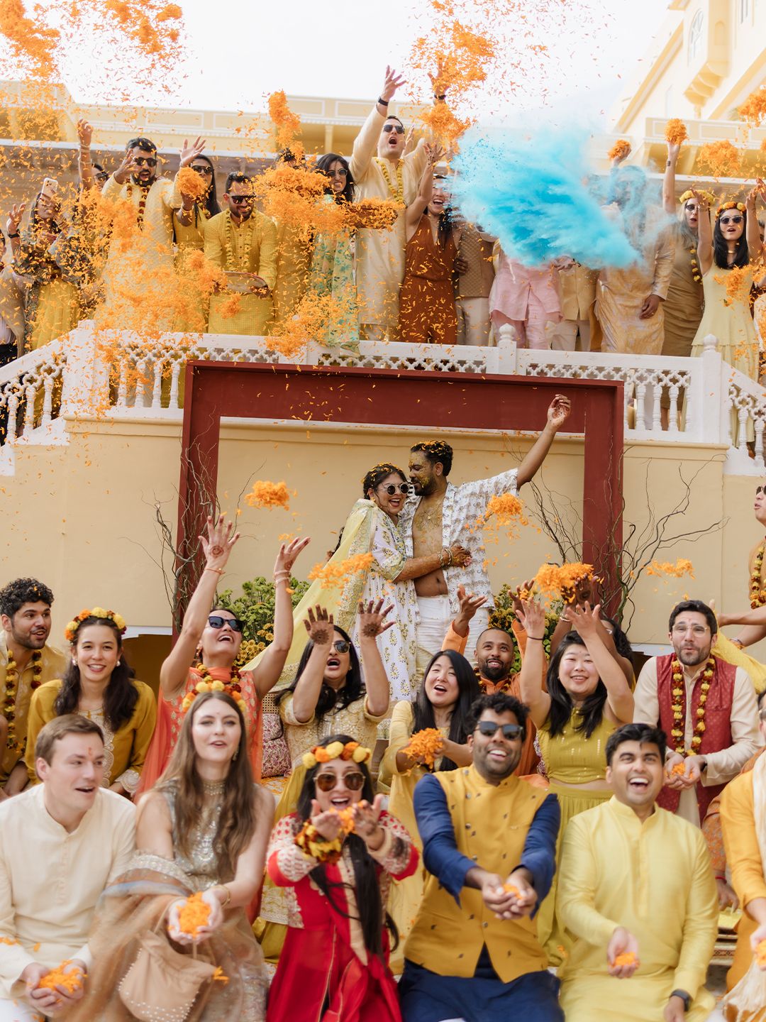 Wedding guests in yellow and orange throw flower petals and colored powder at vibrant haldi celebration.