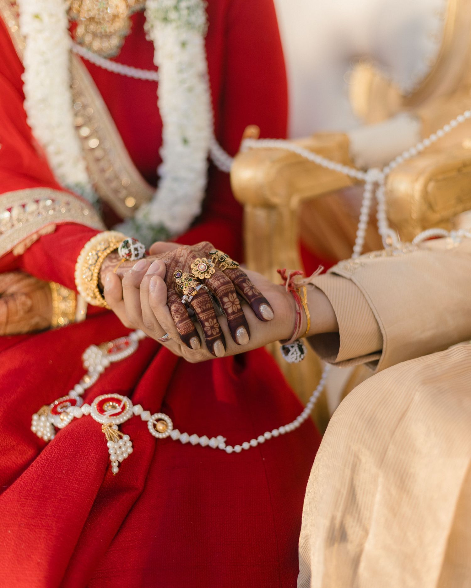 Close-up of Indian bride and groom hands adorned with mehndi, gold jewelry, and pearl accessories.