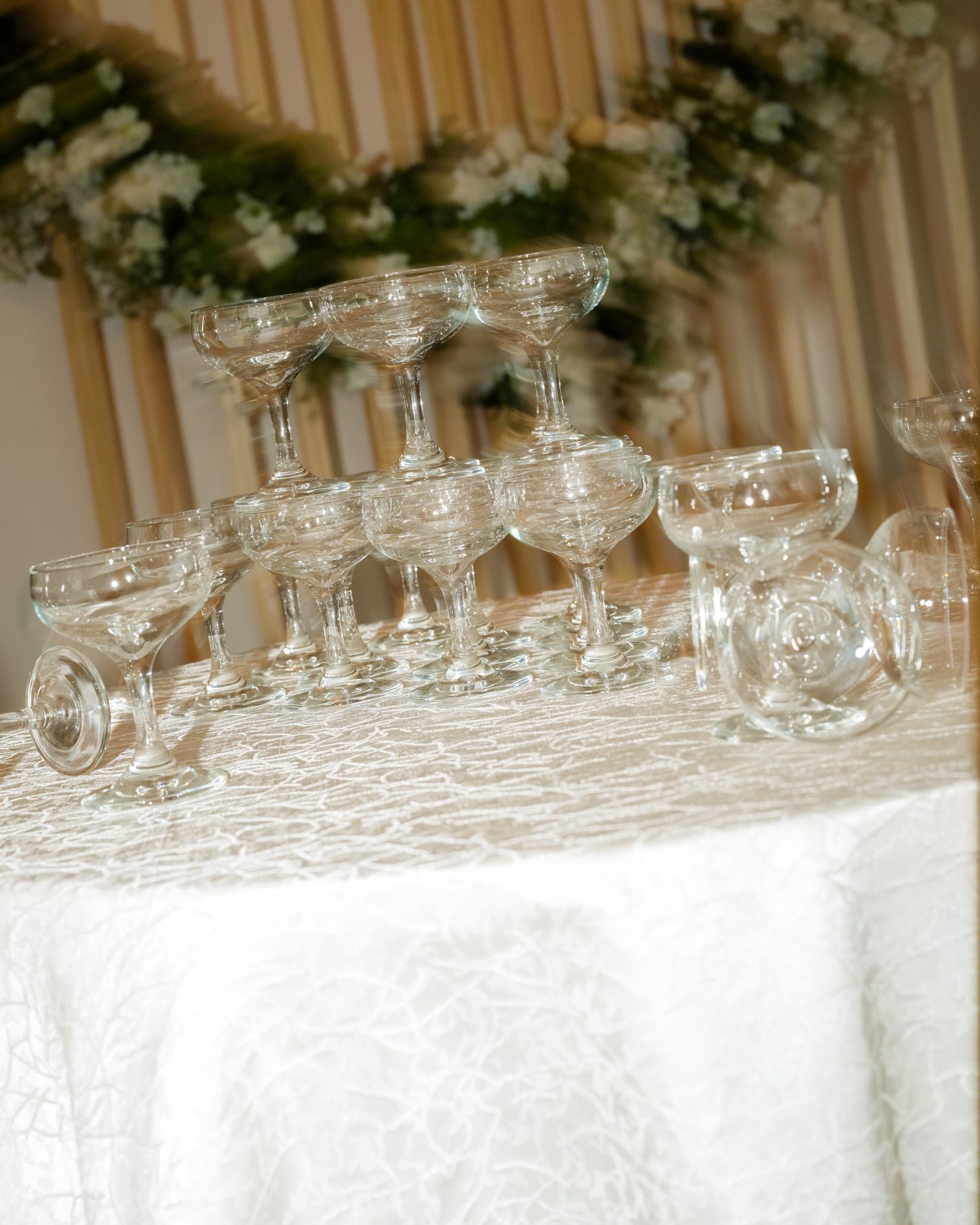 Pyramid of crystal champagne coupes arranged on white linen table with floral backdrop at wedding reception.
