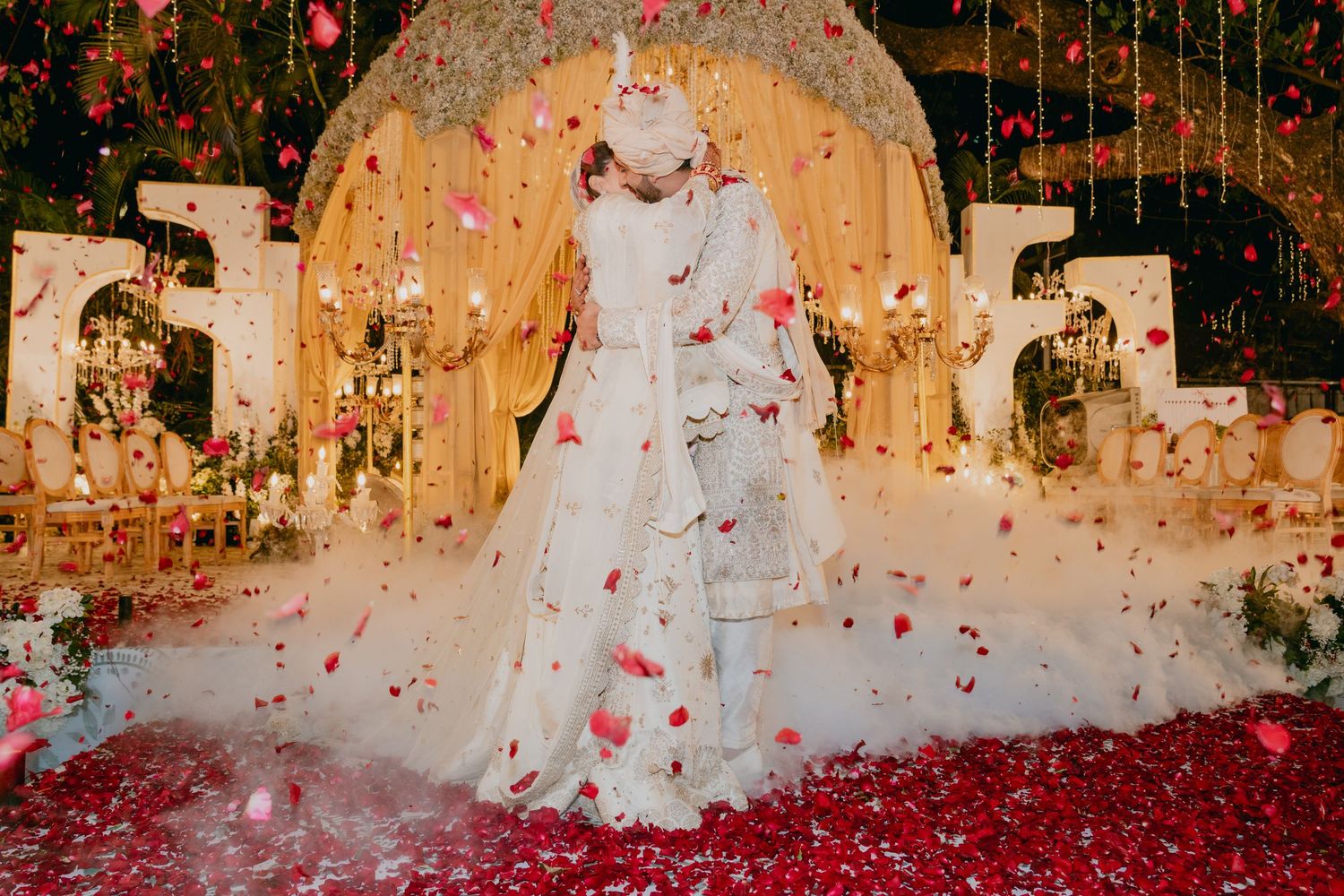 Indian bride and groom share a romantic kiss surrounded by falling rose petals at an elegantly decorated wedding venue.