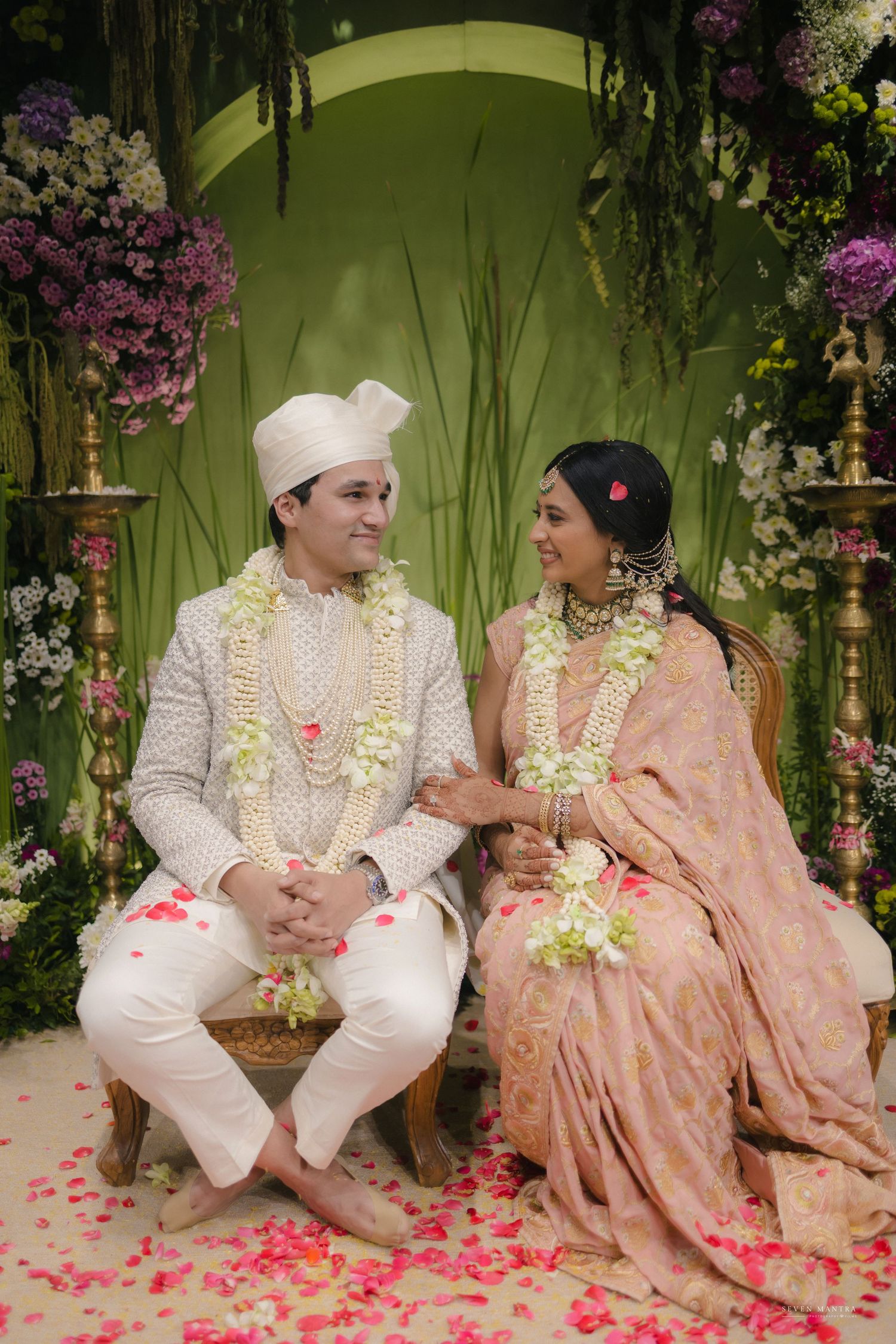 Indian wedding couple in white and pink attire seated with floral garlands at ceremony.