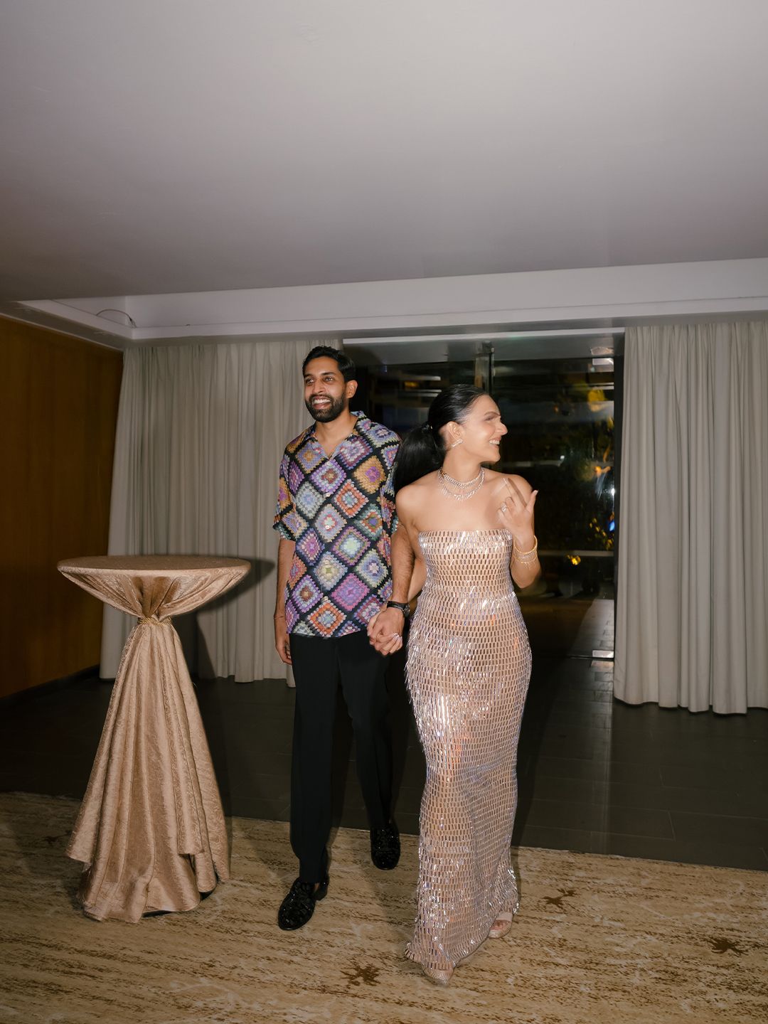 Couple in stylish outfits walk hand in hand at elegant indoor wedding reception venue.