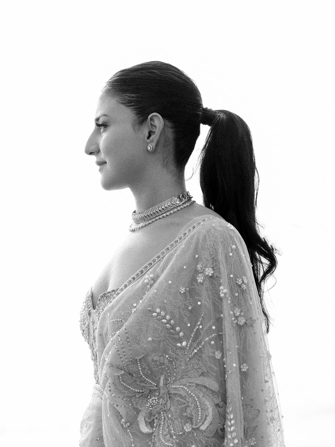 Elegant bride in embroidered saree with pearl necklace and sleek ponytail in black and white portrait.