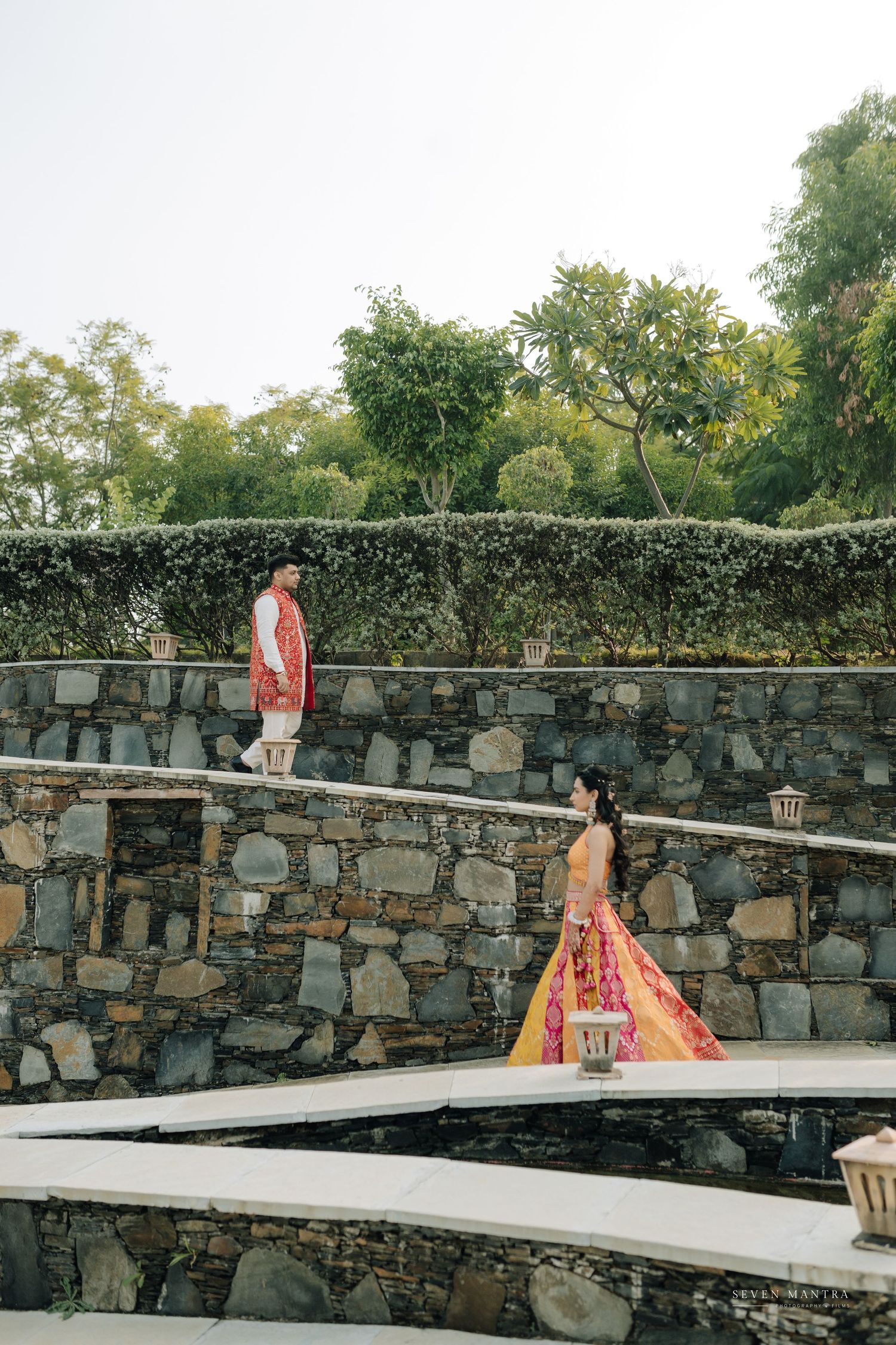Indian bride and groom in traditional attire walk separately along stone terrace during wedding photoshoot.