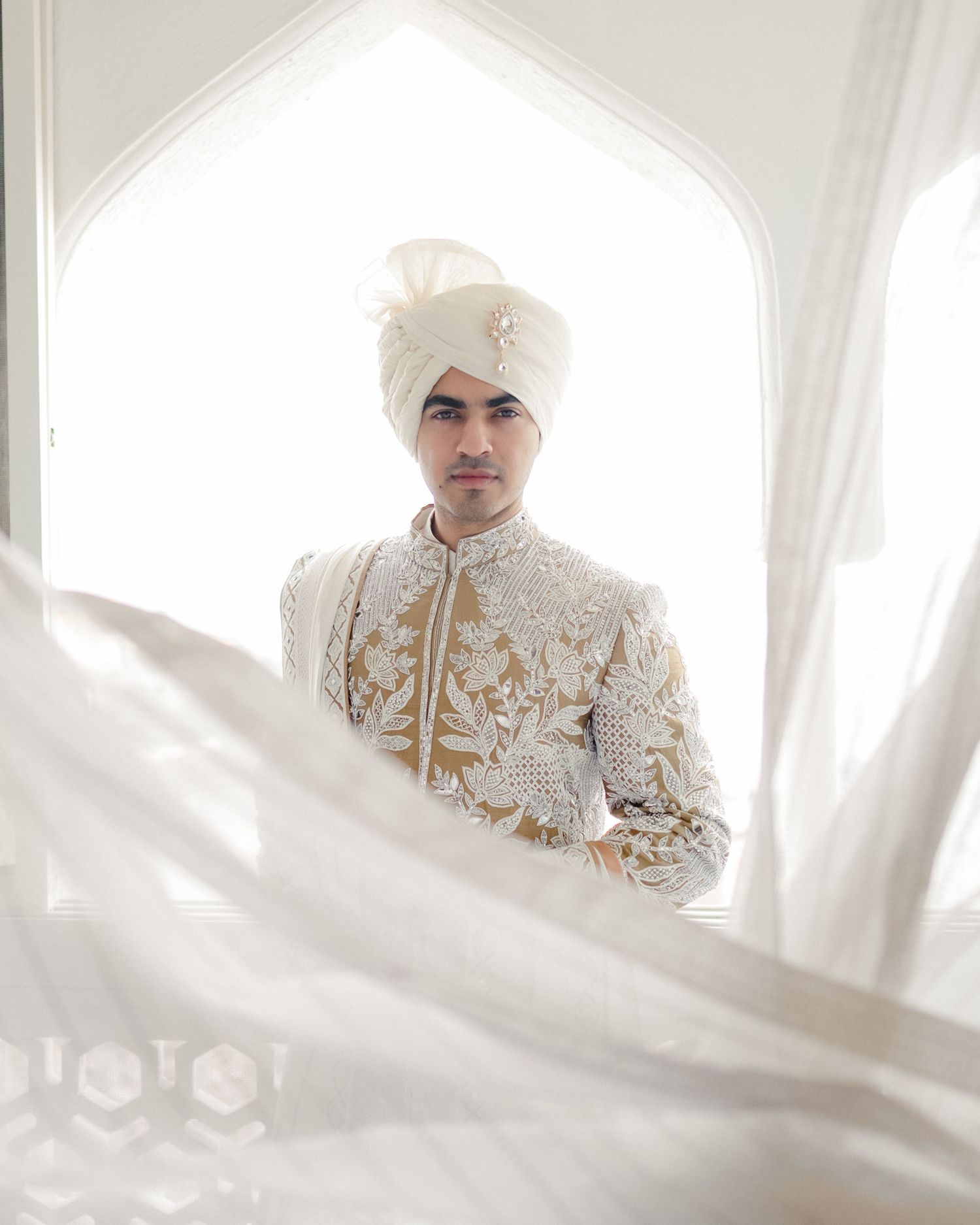 Indian groom in ivory embroidered sherwani and white turban standing in elegant arched doorway.