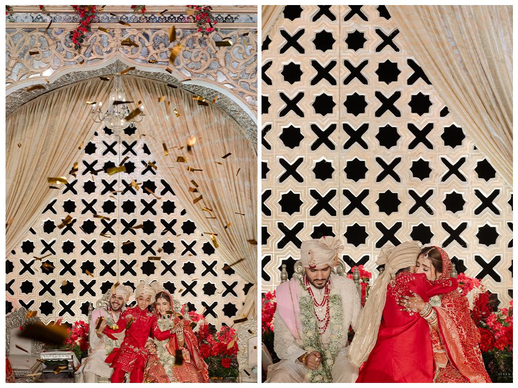 Indian wedding ceremony couple in red bridal outfits seated under ornate white lattice backdrop with golden drapes.