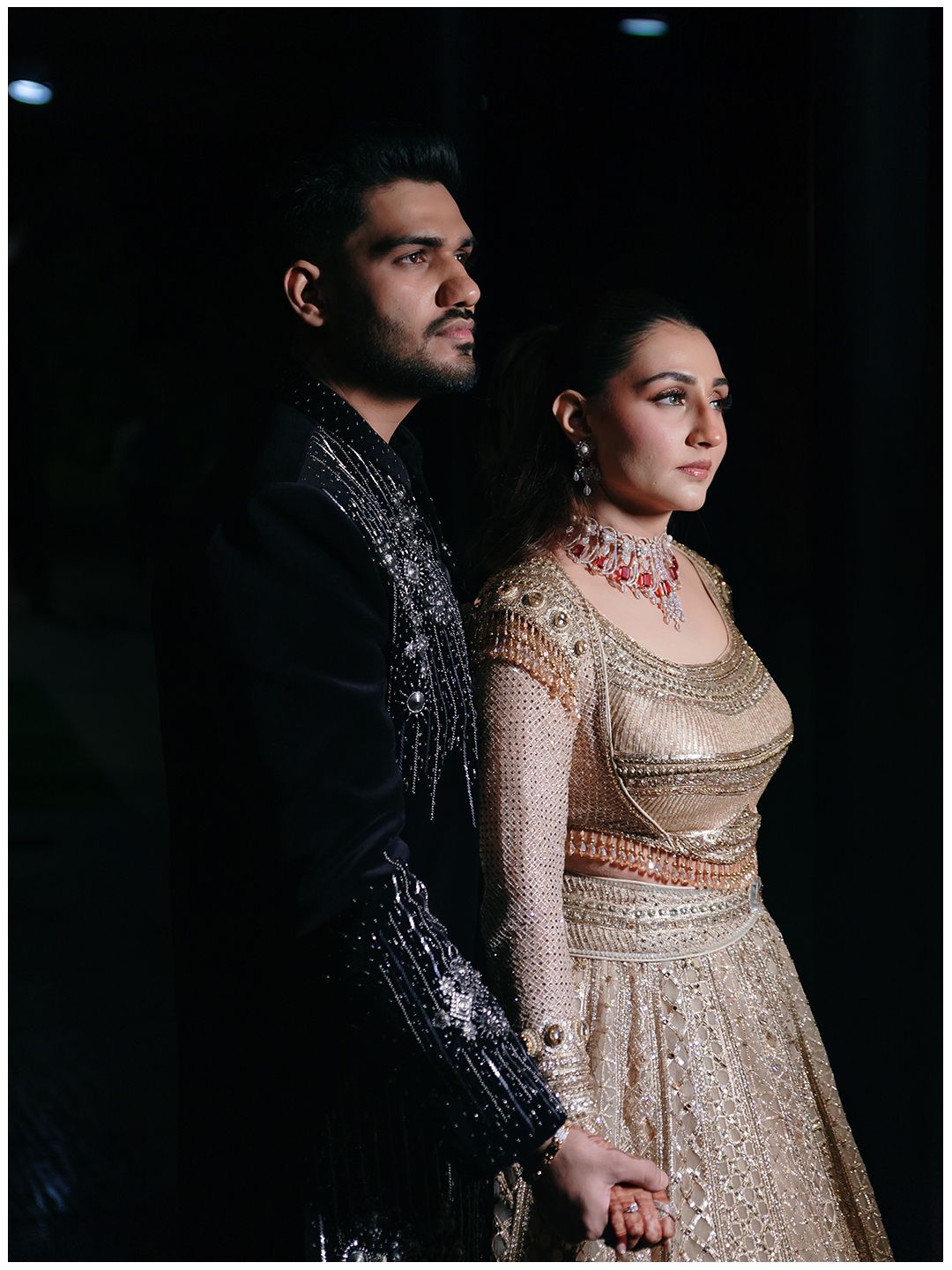 Elegant Indian couple in gold lehenga and black embroidered sherwani posing in dramatic dark studio lighting.