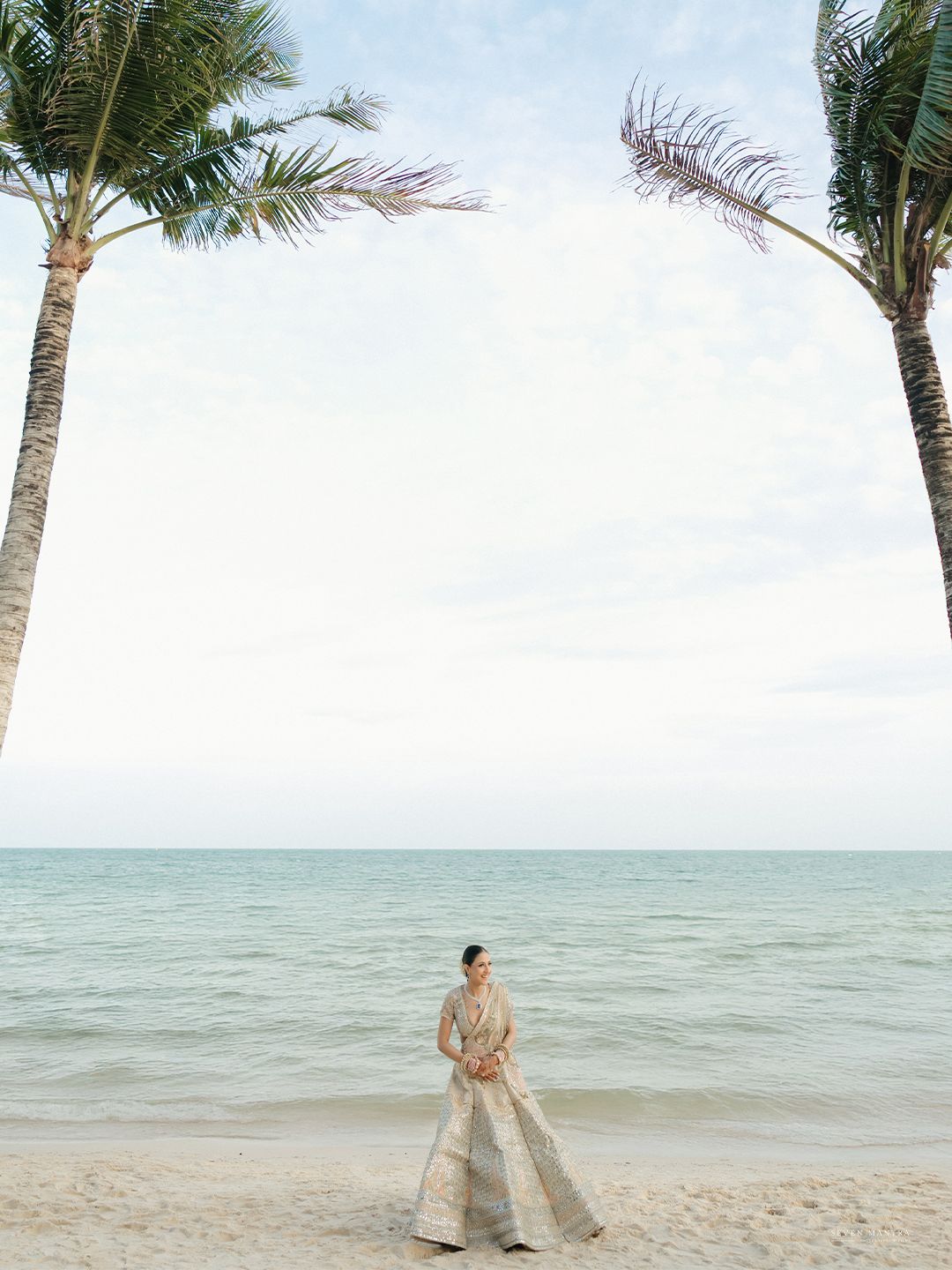 Indian bride in embroidered lehenga stands gracefully between two palm trees on a tropical beach.