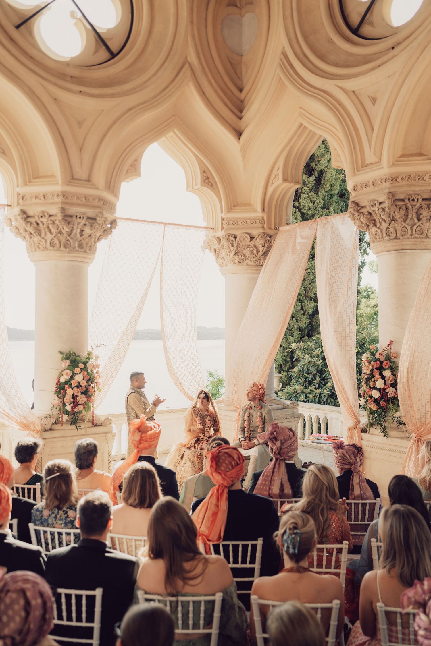 Indian wedding ceremony inside ornate arched pavilion with white draped mandap and guests seated on white chairs.