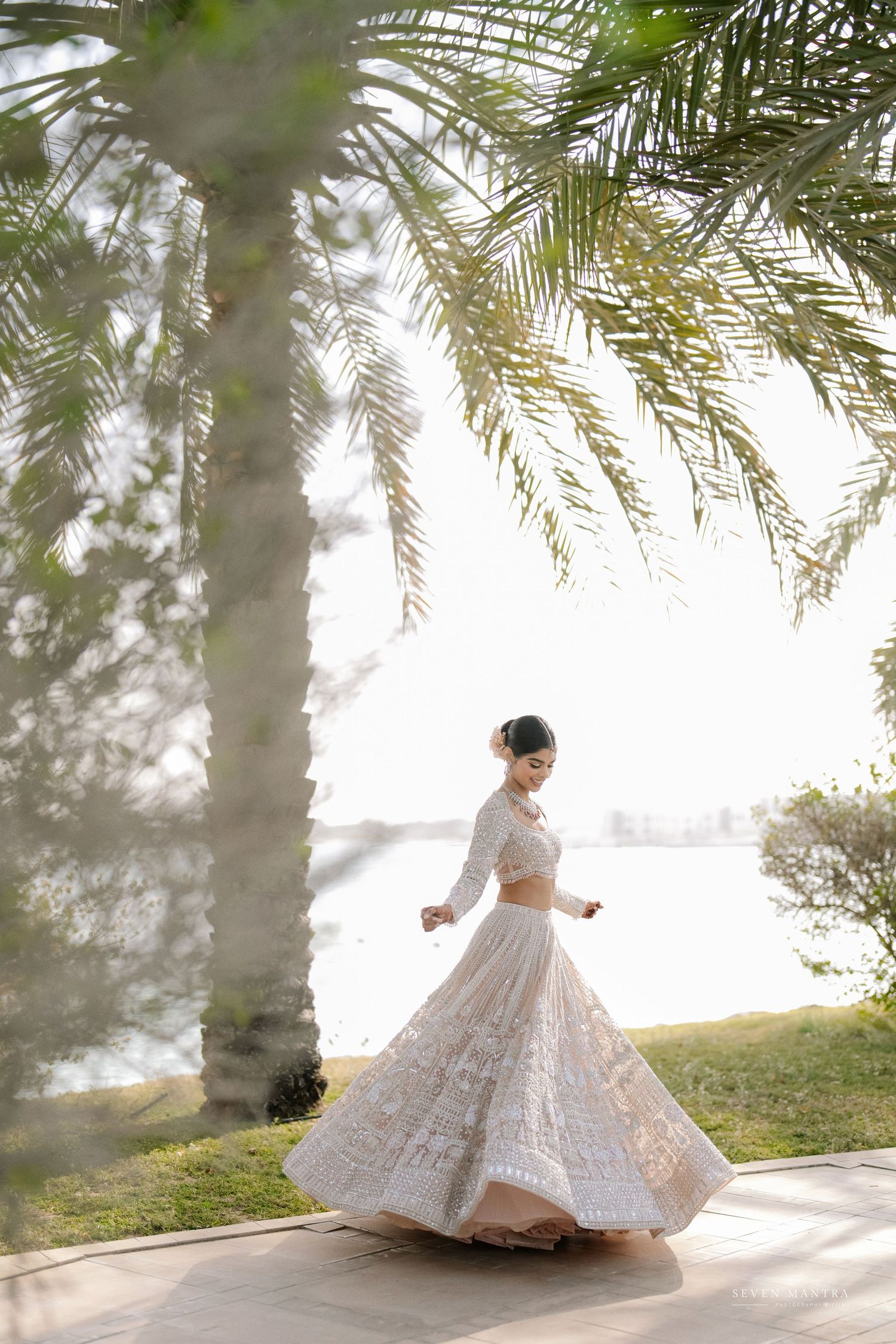 Bride twirling in white embroidered lehenga under palm trees beside a serene waterfront backdrop.