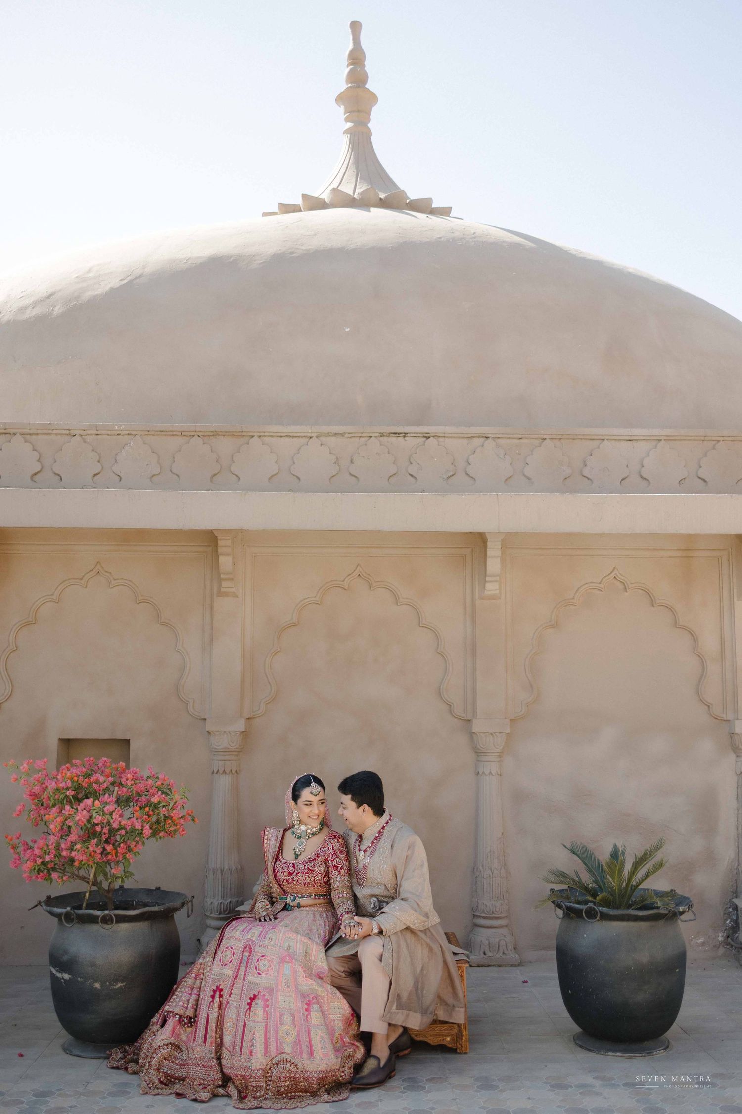 Bride and groom in traditional Indian wedding attire seated before an ornate Rajasthani dome architecture.