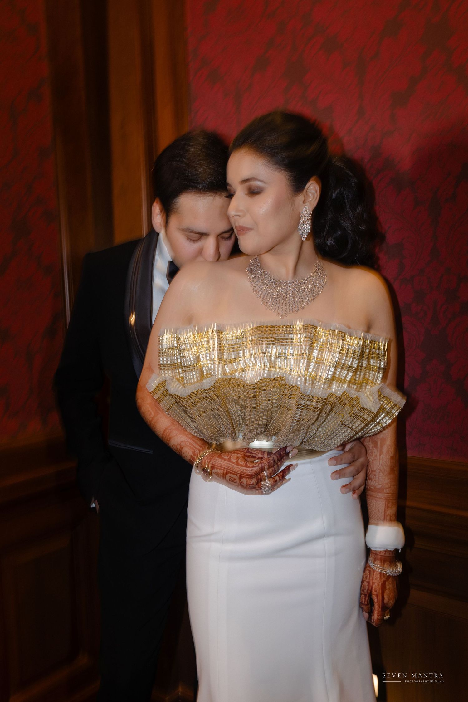 Bride in white gown with gold fan accessory and mehndi, groom in tuxedo sharing intimate moment.