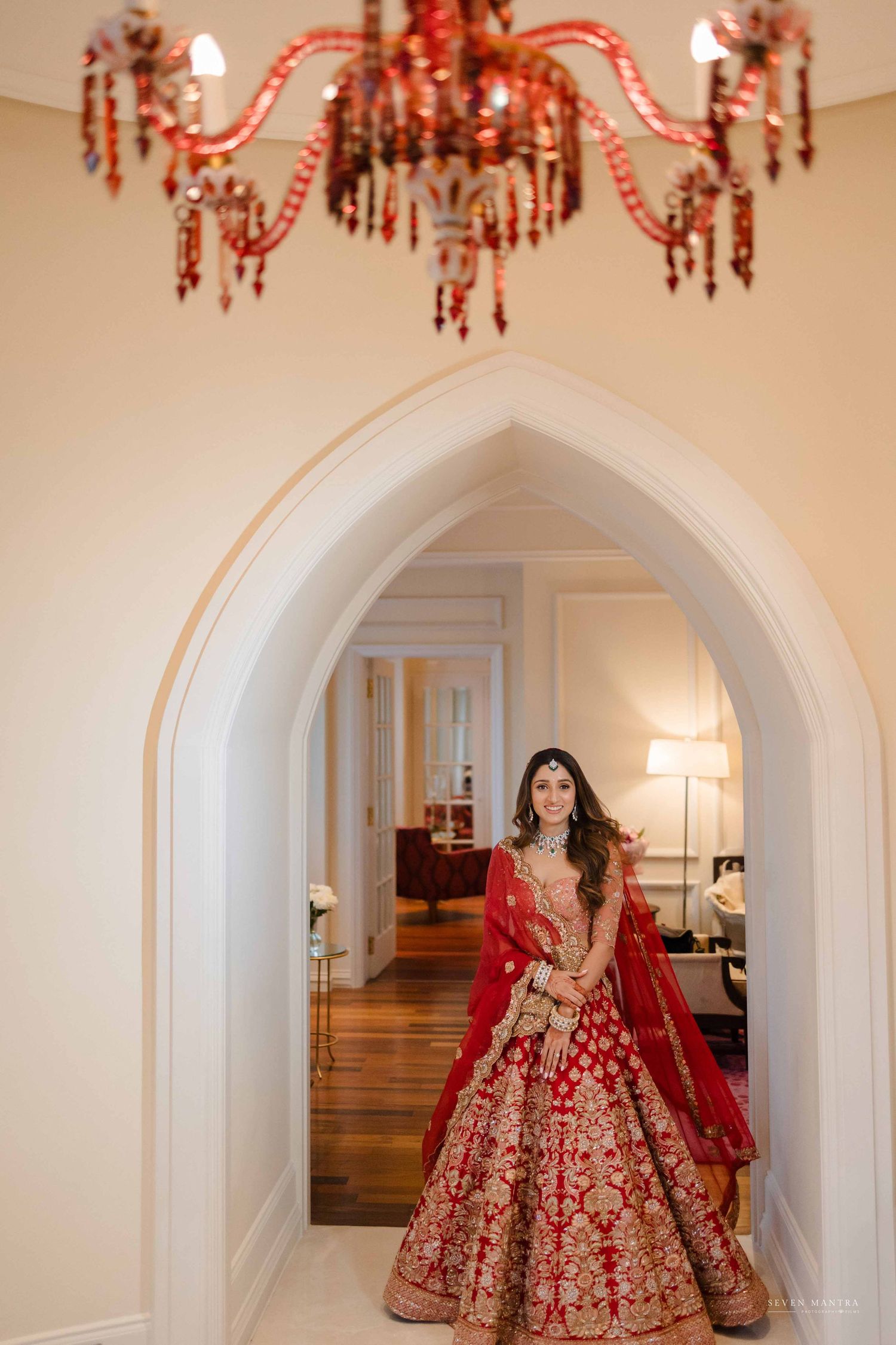 Indian bride in red and gold lehenga stands under ornate chandelier in arched doorway.