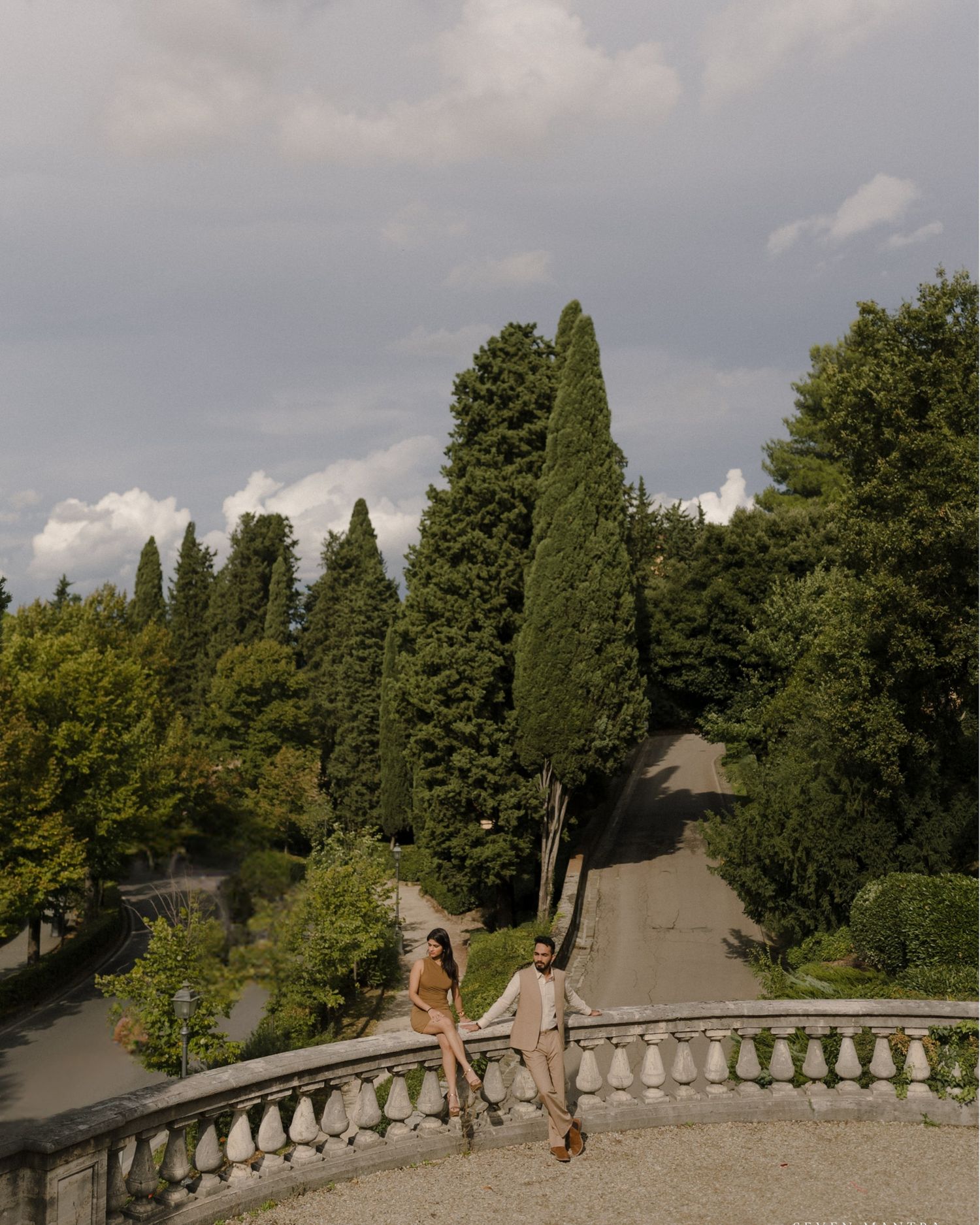 Couple posing on a stone balustrade terrace surrounded by tall cypress trees in an Italian garden.