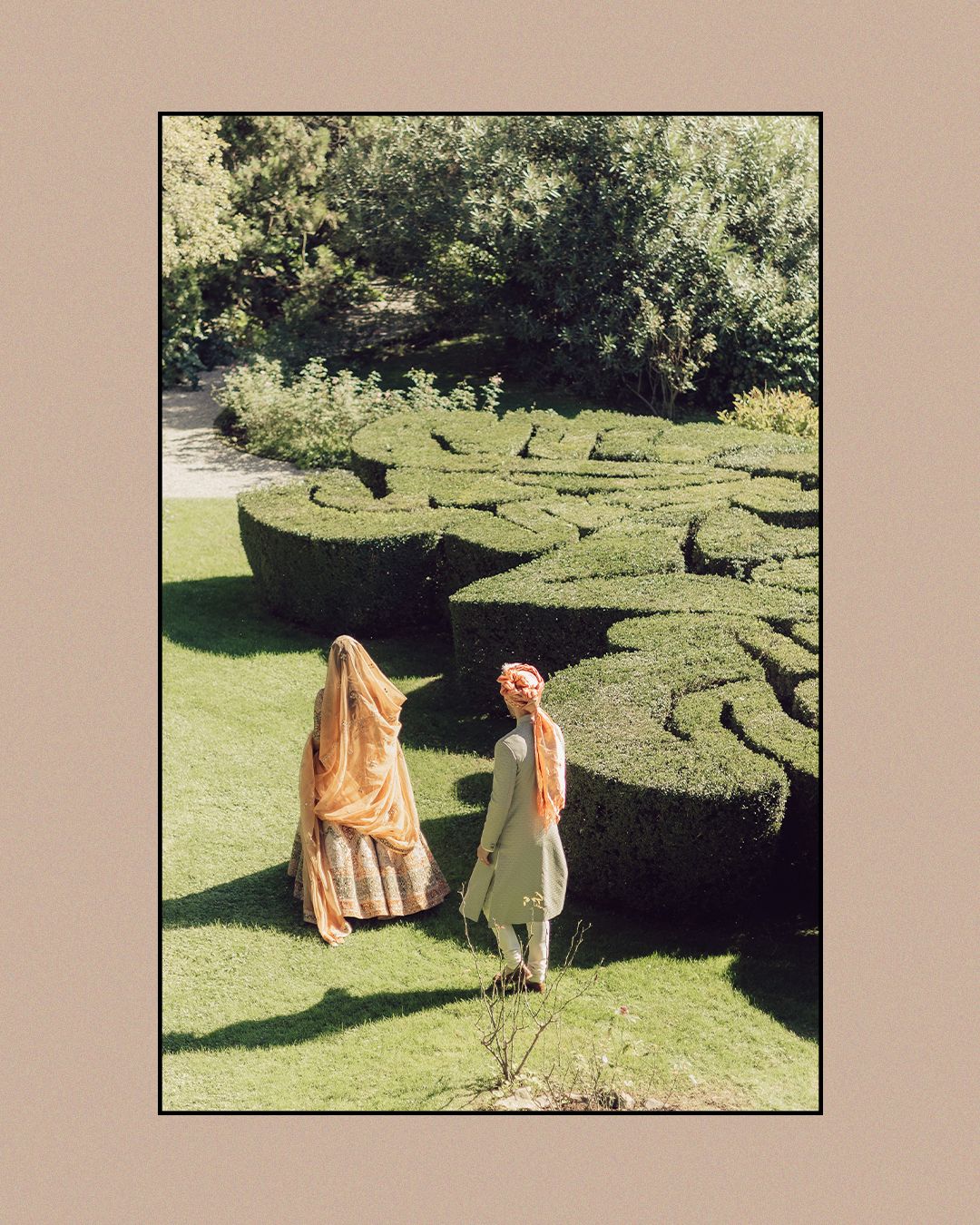 Bride in orange lehenga with dupatta veil and groom in kurta explore sculpted topiary garden maze.