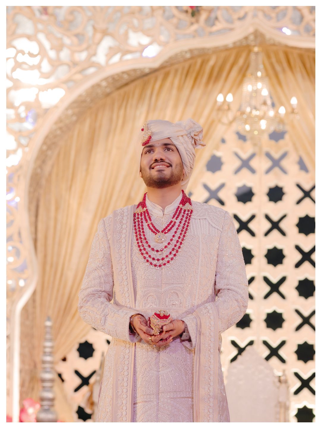 Handsome Indian groom in cream embroidered sherwani with red floral garland at wedding mandap.