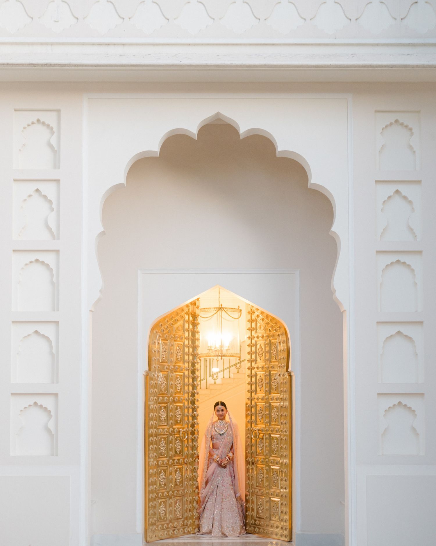 Indian bride in blush pink lehenga standing in an ornate golden arched doorway of a white palace.