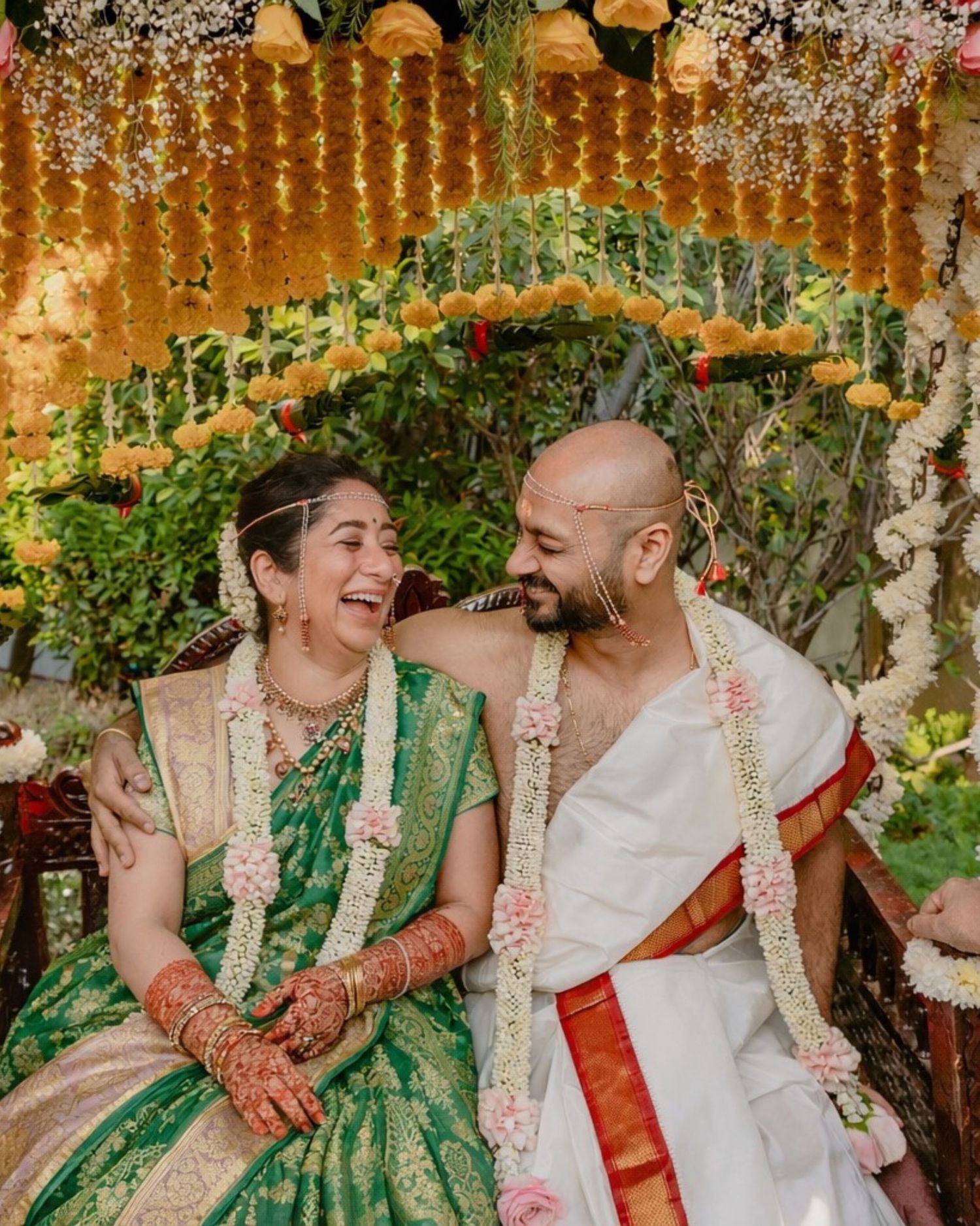Joyful newlyweds in green silk saree and white dhoti laughing under floral mandap at South Indian wedding.