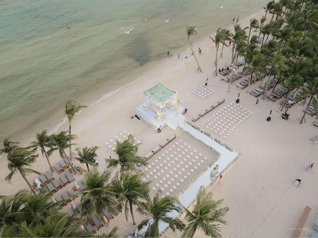 Aerial view of a beachfront Indian wedding mandap setup with white chairs on tropical sandy beach.