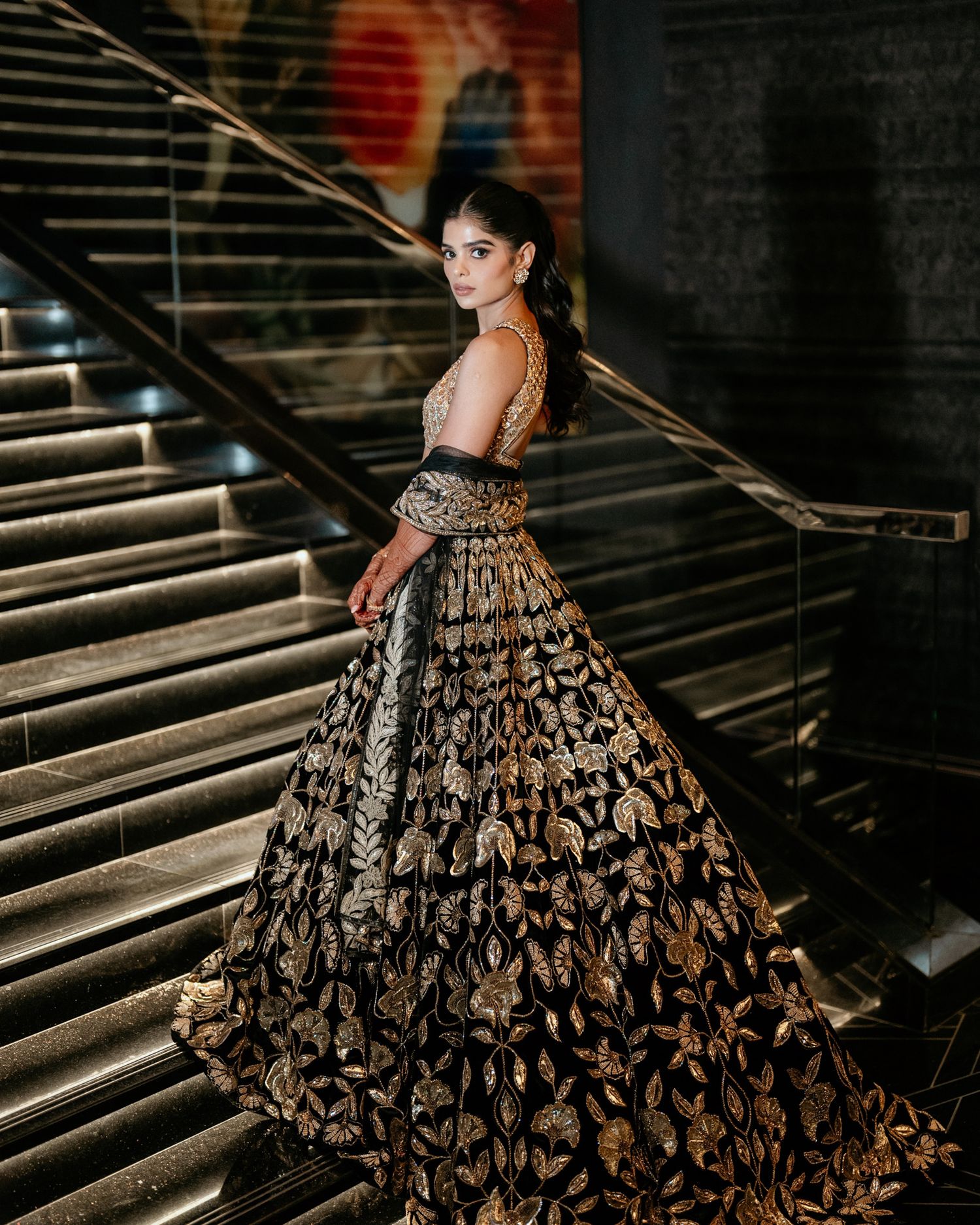 Bride in stunning black and gold embroidered lehenga posing elegantly on a grand staircase.