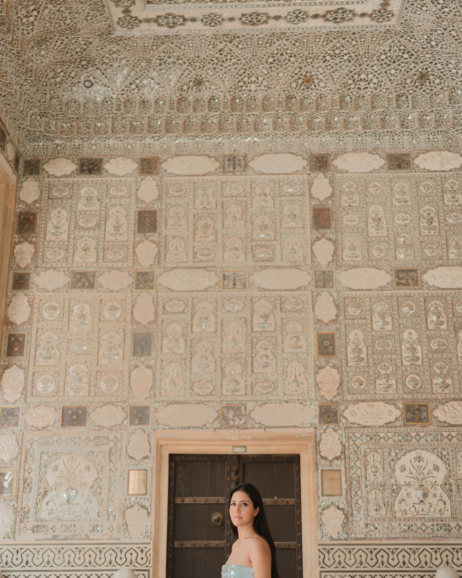 Woman in a light blue dress standing beneath an elaborately decorated Mughal-style mirrored ceiling in a historic palace.