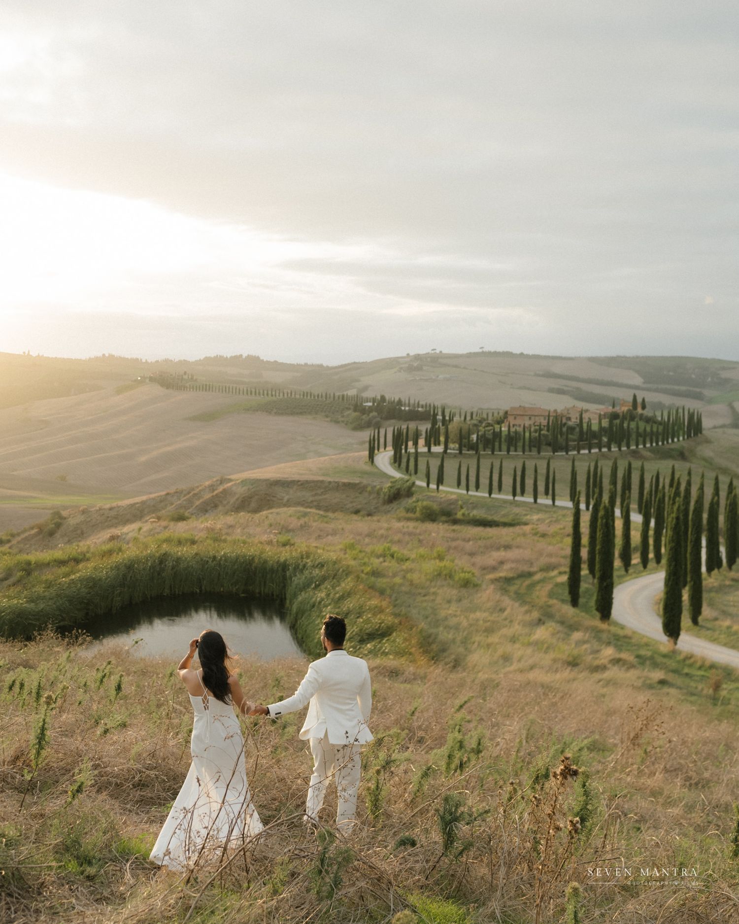 Wedding couple holding hands overlooking rolling Tuscan hills with cypress trees at golden sunset.
