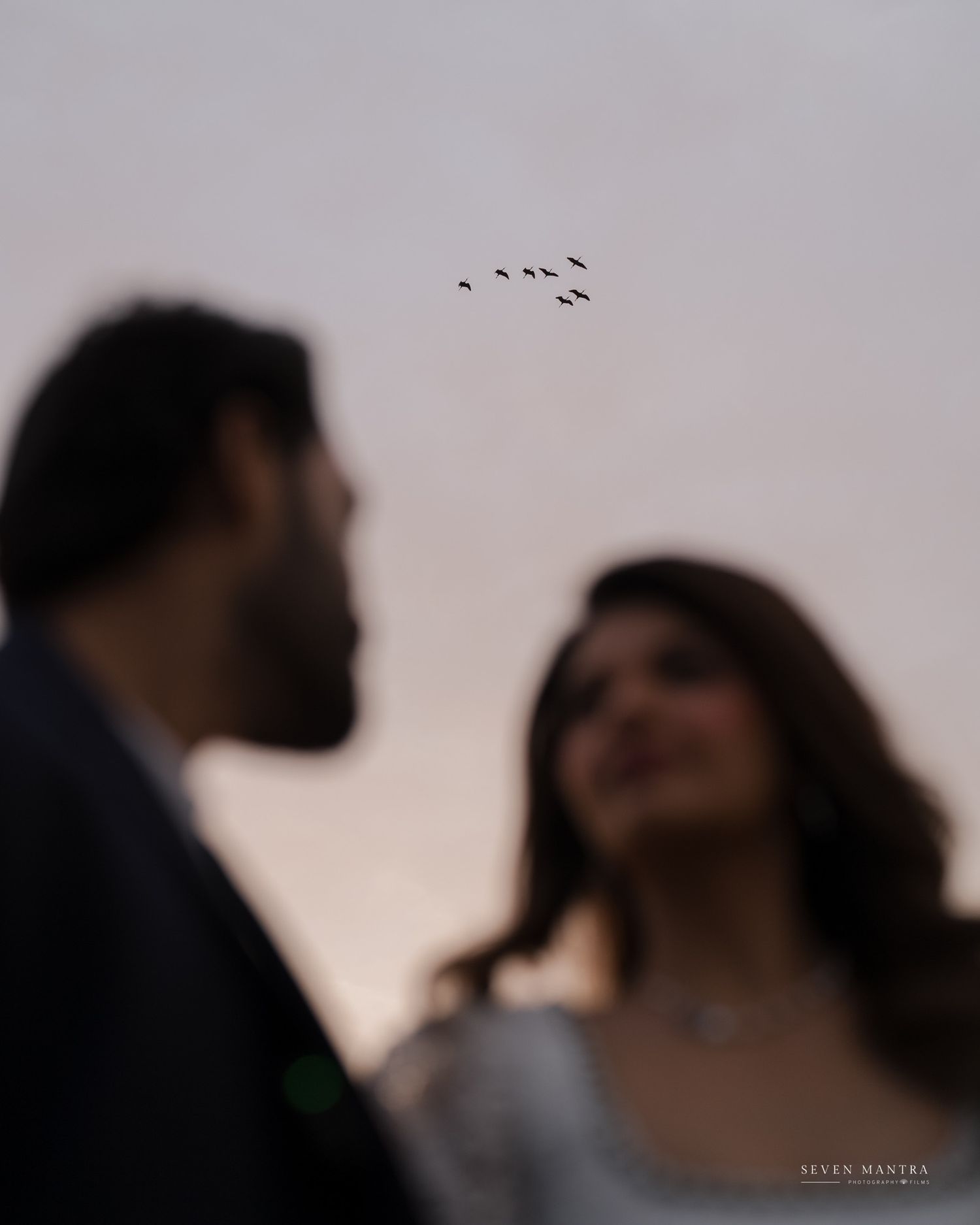 Romantic blurred couple portrait with birds flying in sky, woman in blue outfit smiling.