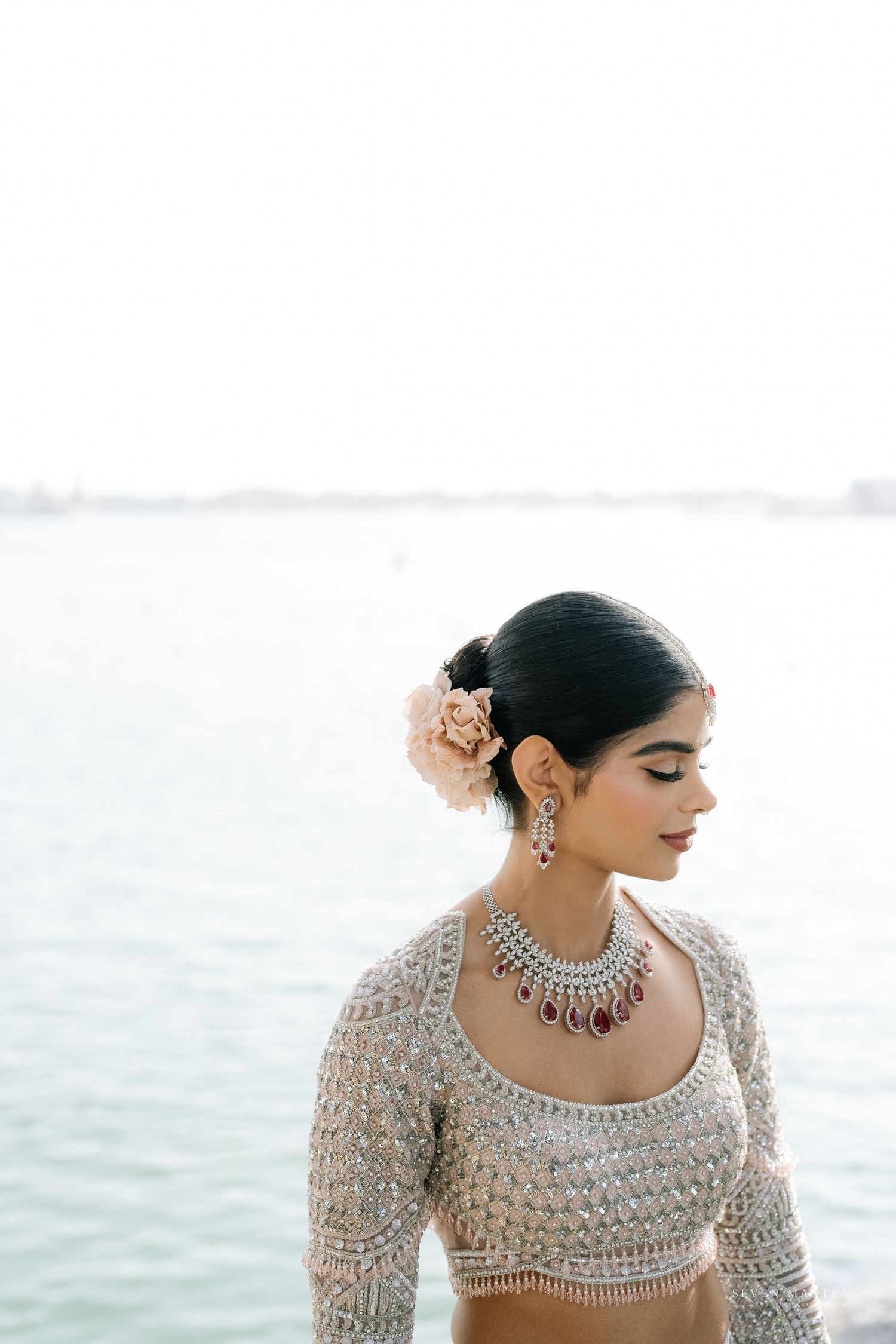 Elegant Indian bride in silver embroidered blouse with floral hair accessory and ruby necklace by water.