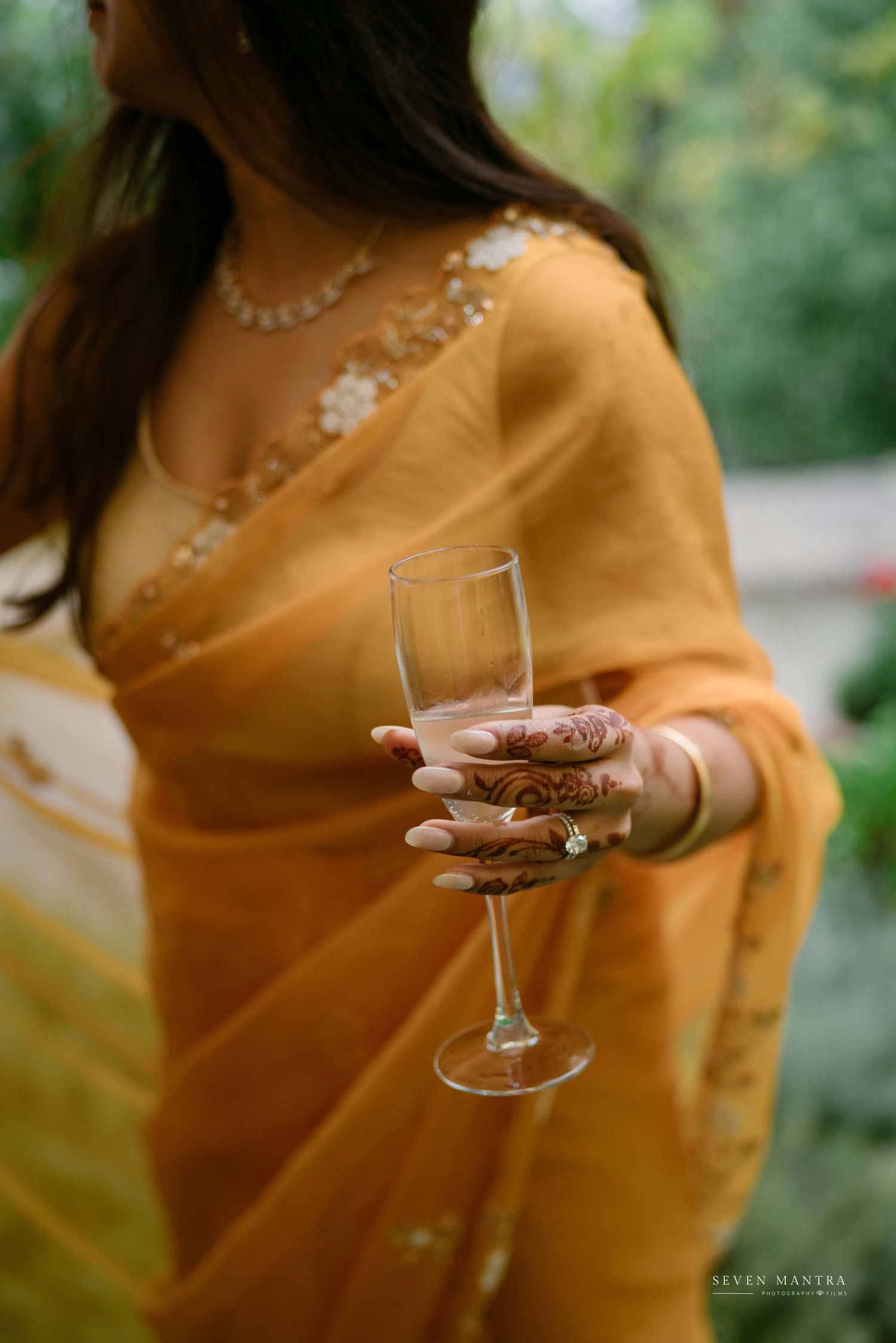 Bride in yellow saree with mehndi hands elegantly holding a champagne glass.