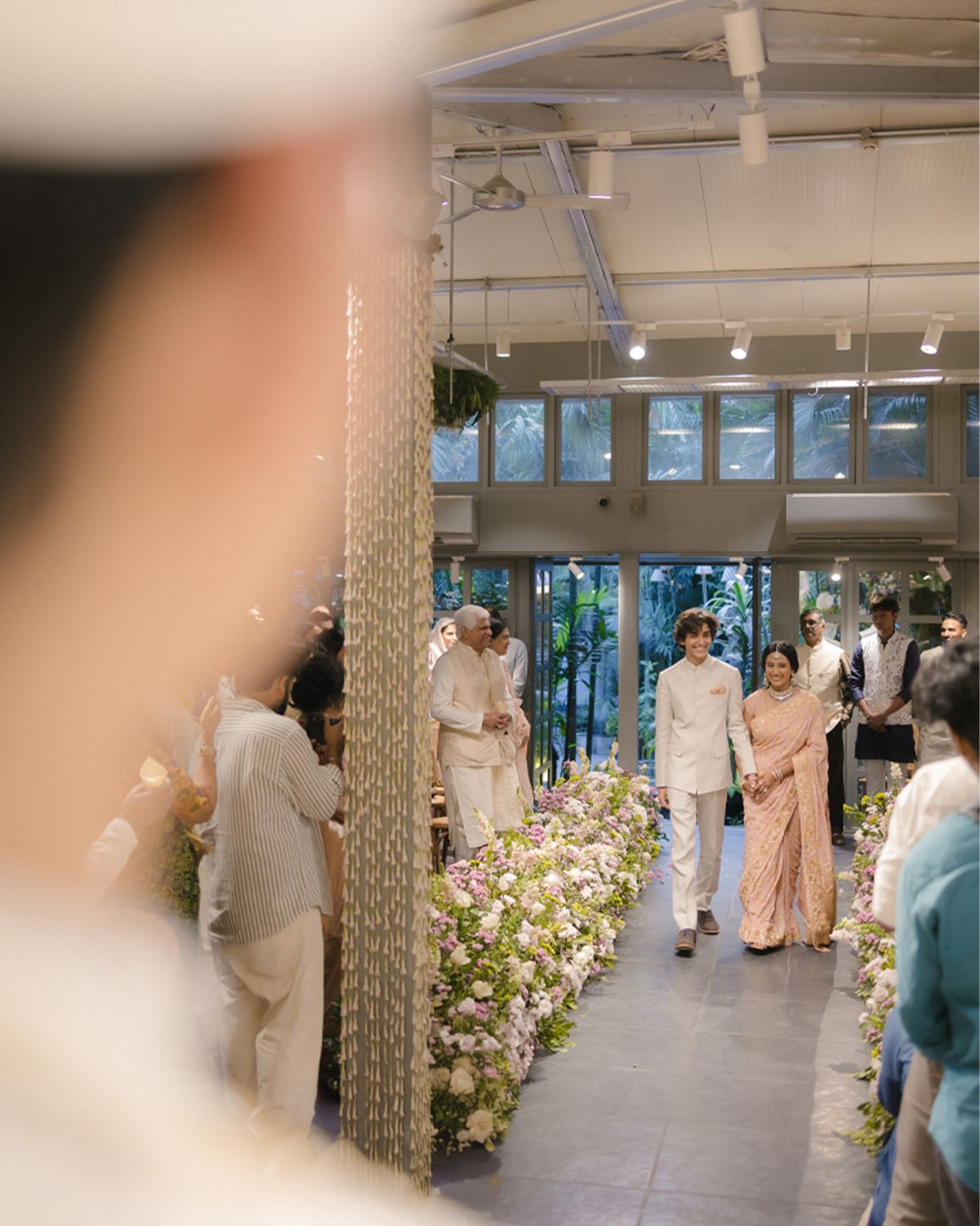 Groom awaiting bride walking down floral-lined aisle in modern Indian wedding venue.