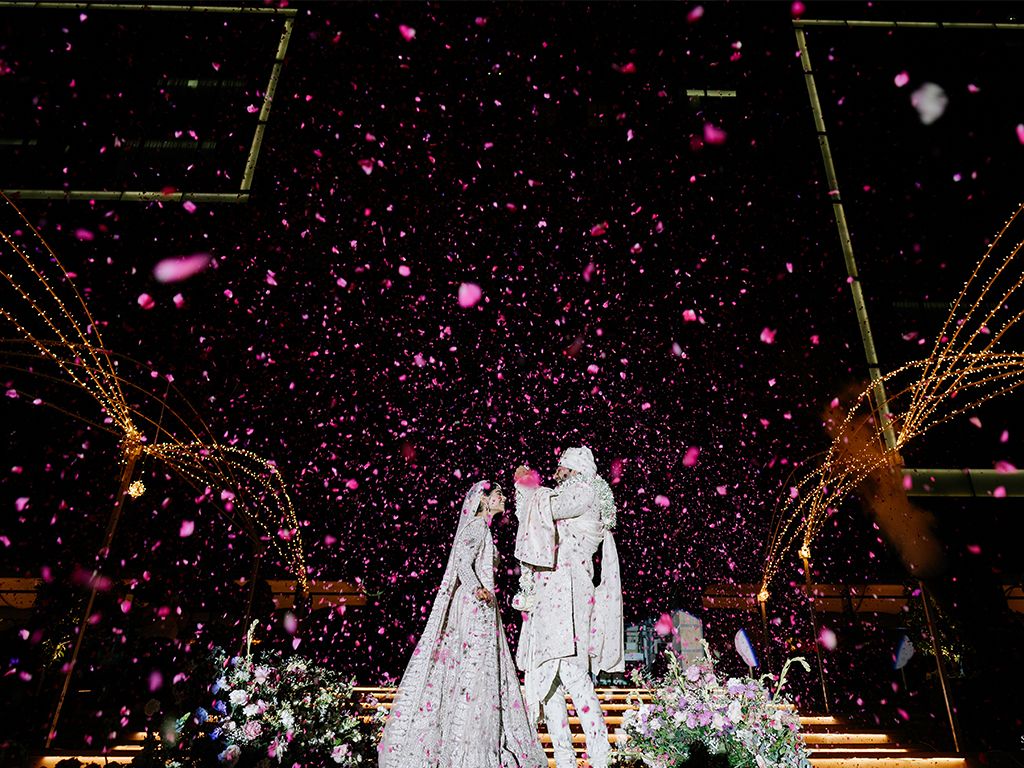 Indian bride and groom stand beneath shower of pink confetti petals at spectacular nighttime wedding celebration.