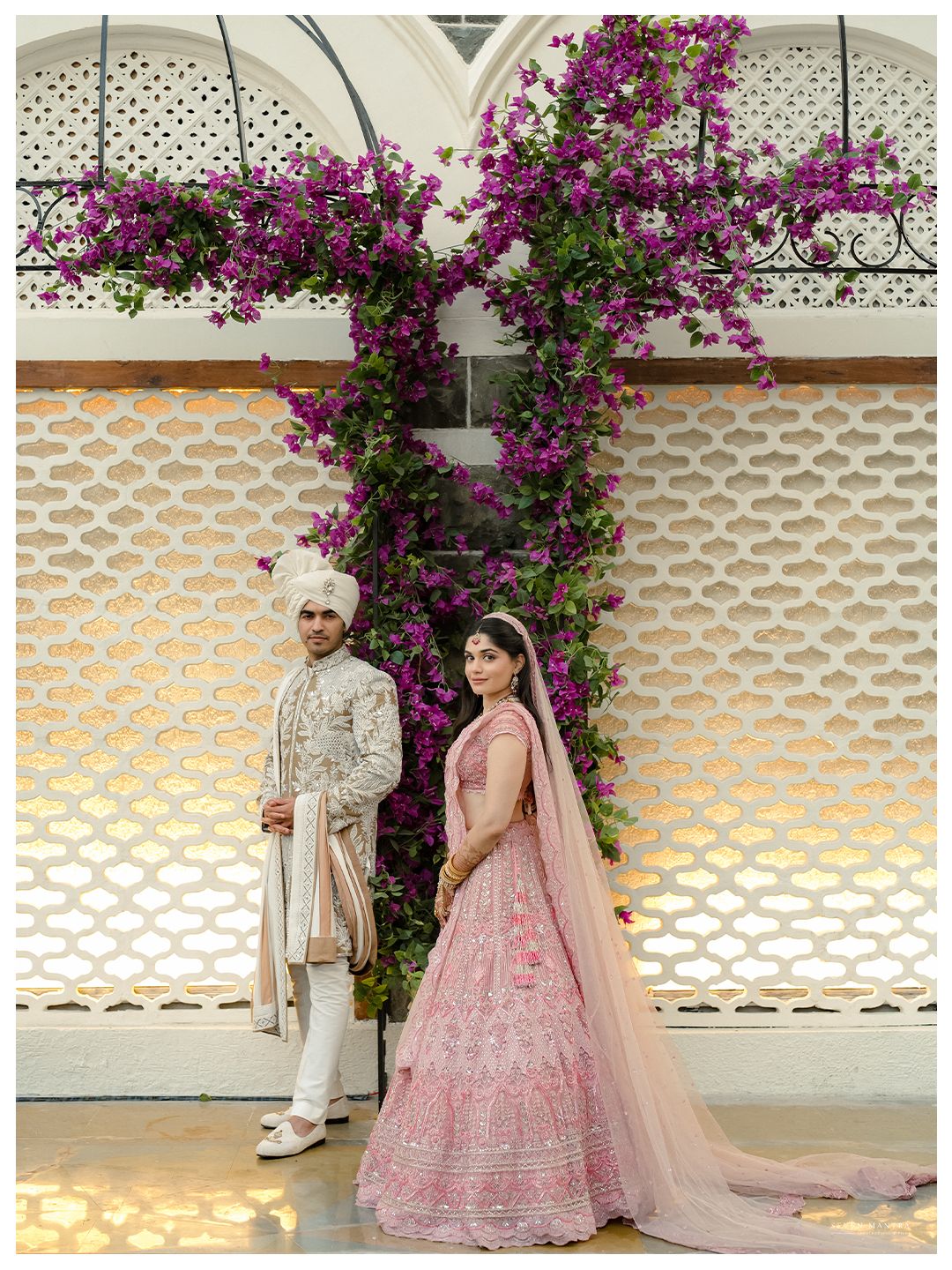 Indian wedding couple posing before ornate white wall with vibrant purple bougainvillea flowers in Mumbai.