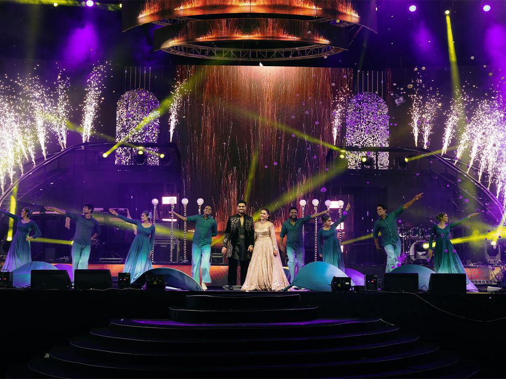 Grand wedding sangeet performance on stage with dancers in teal outfits and spectacular purple light and fireworks show.