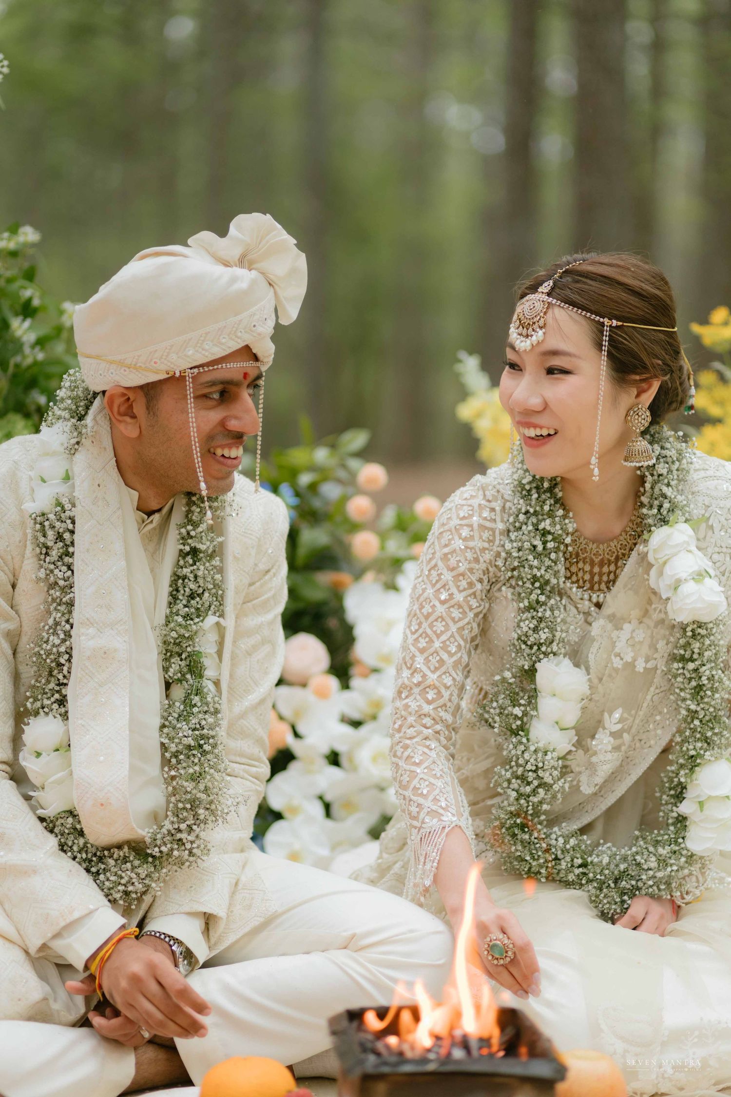 Smiling couple in white wedding attire exchanging glances during Hindu fire ritual in forest.
