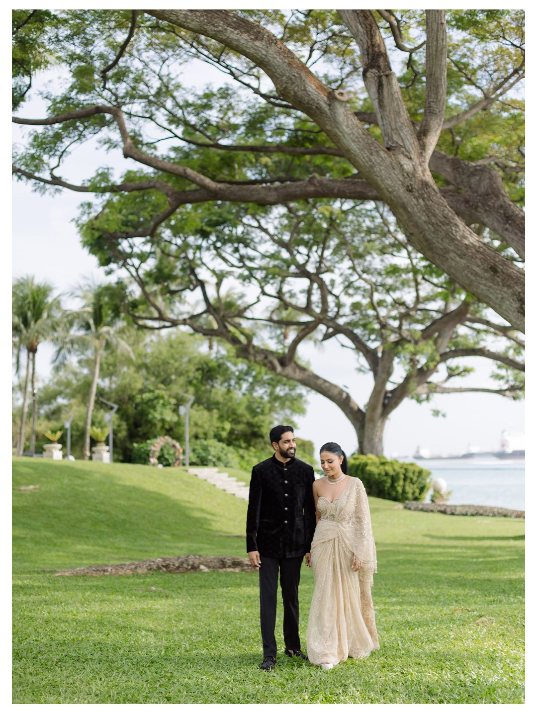 Couple in traditional Indian wedding attire walking on lush green lawn by the waterfront.