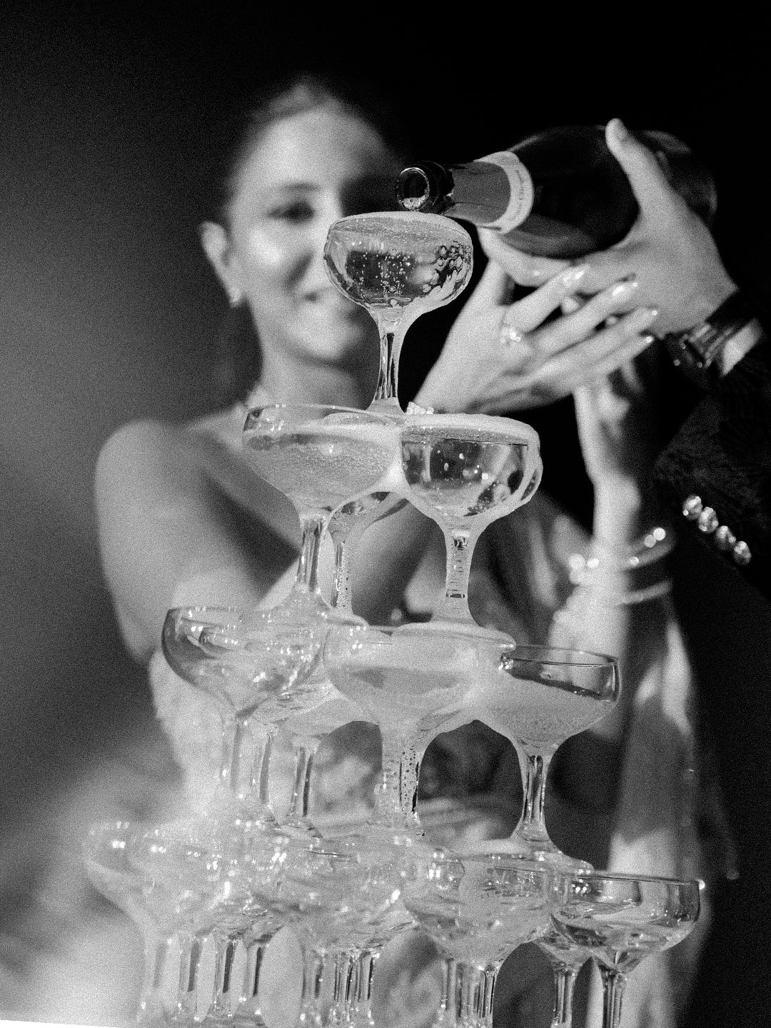 Black and white photo of bride and groom pouring champagne into a pyramid of glasses at wedding.