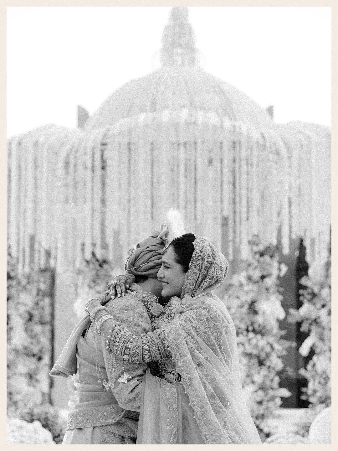 Black and white photo of Indian bride and groom embracing under ornate floral dome at wedding ceremony.