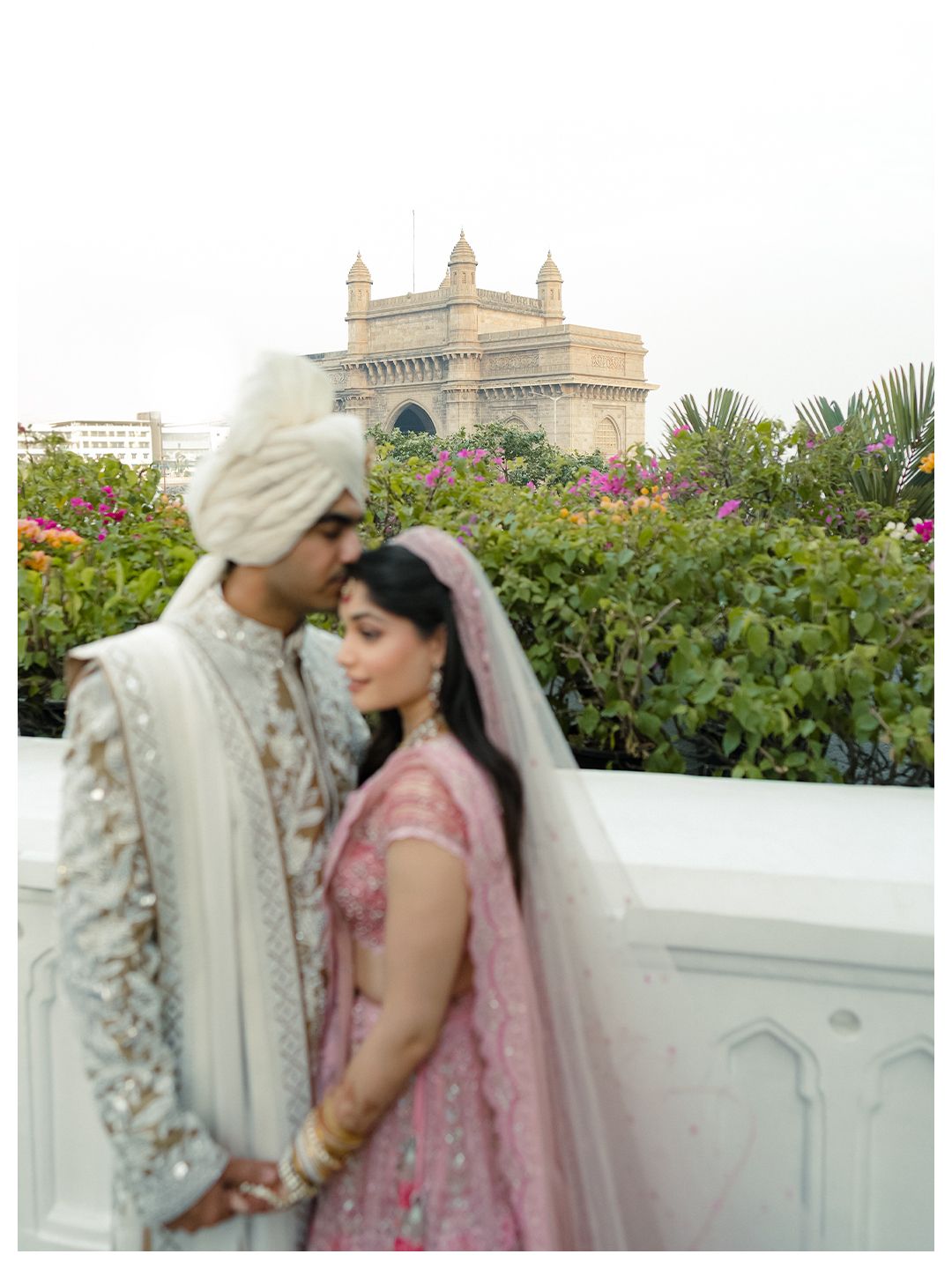 Indian wedding couple in white sherwani and pink lehenga posing before the Gateway of India in Mumbai.