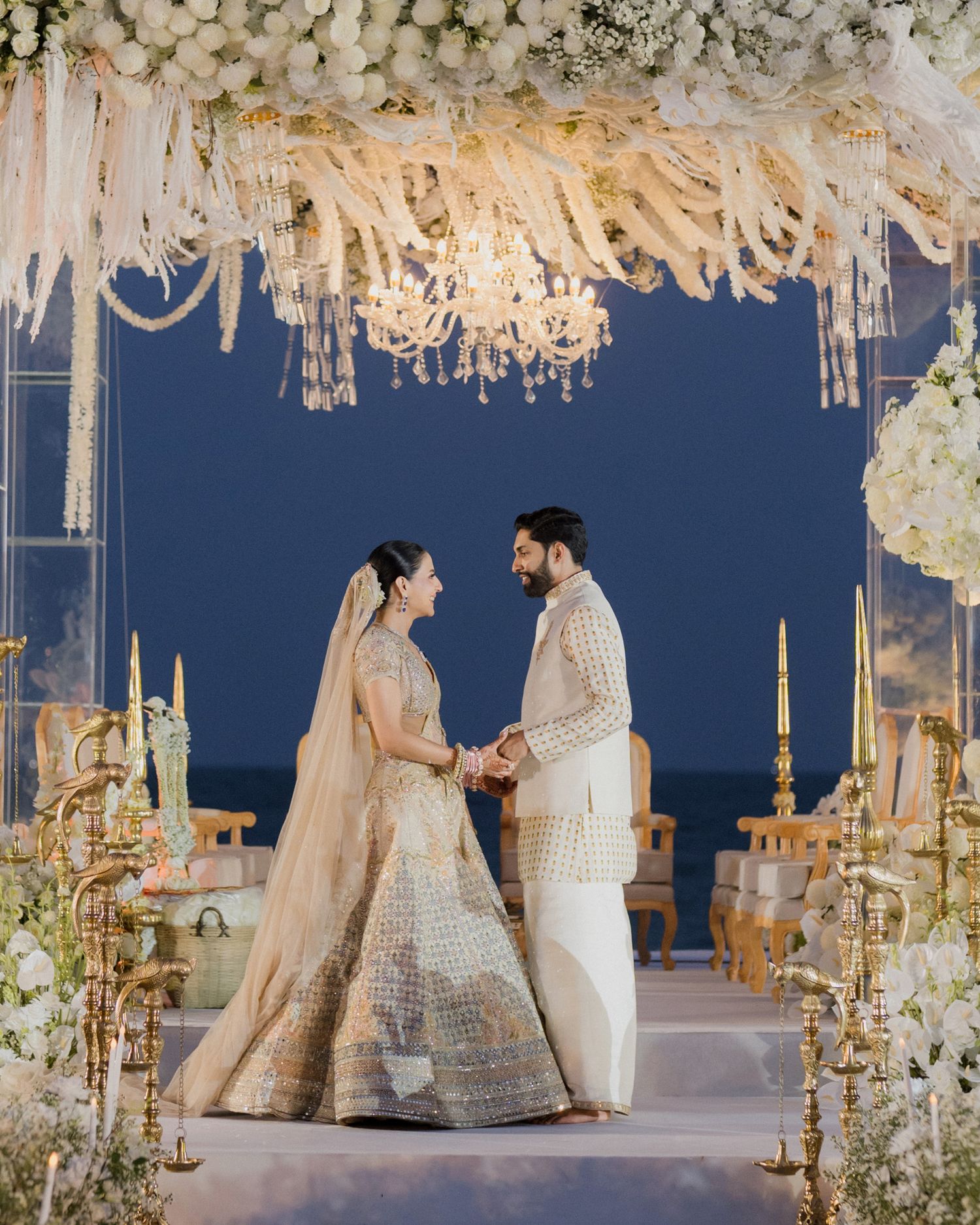 Bride and groom exchange vows under floral chandelier mandap at elegant beachfront wedding ceremony.